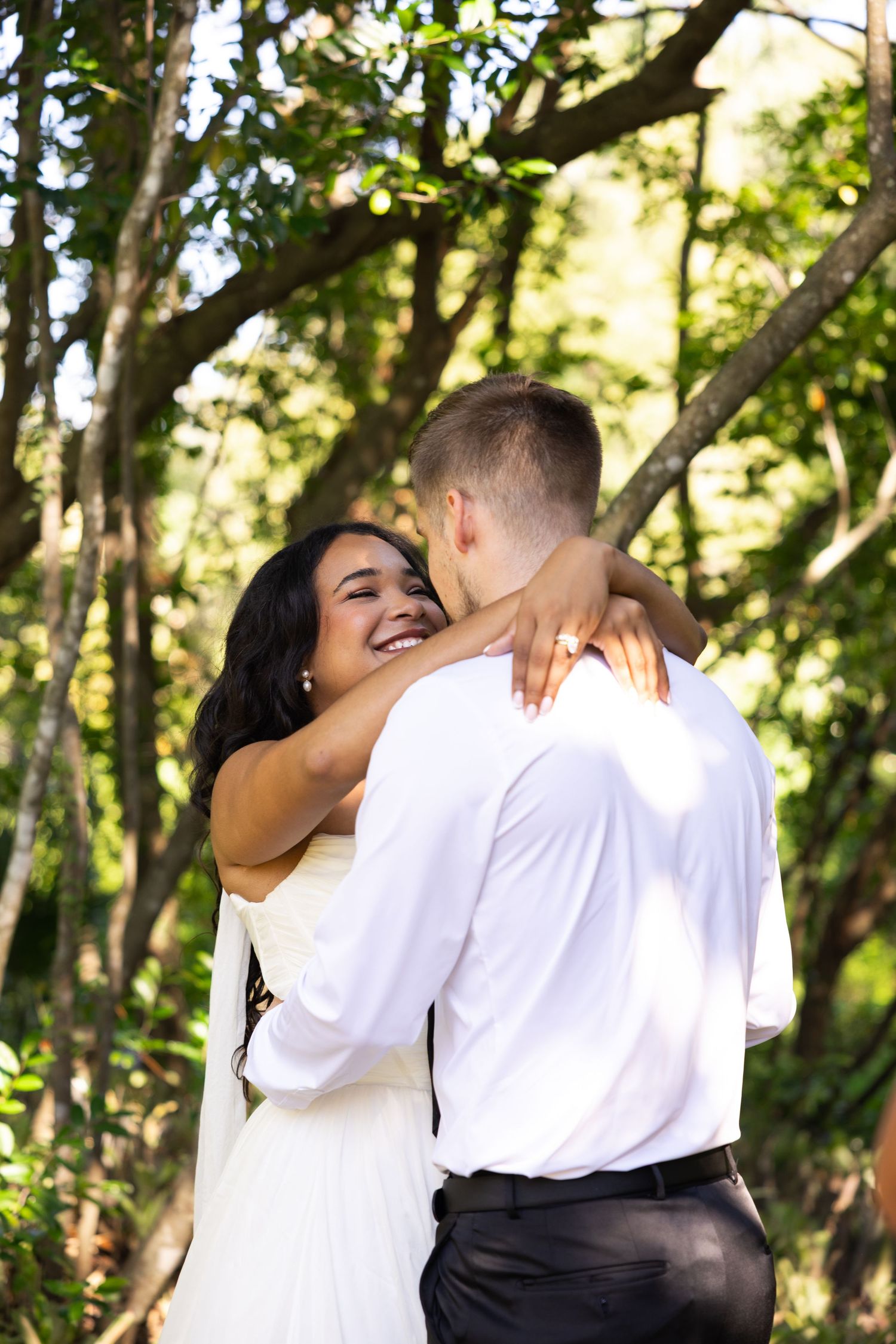 A joyful bride in a white dress embraces her groom outdoors, smiling radiantly among lush green trees.