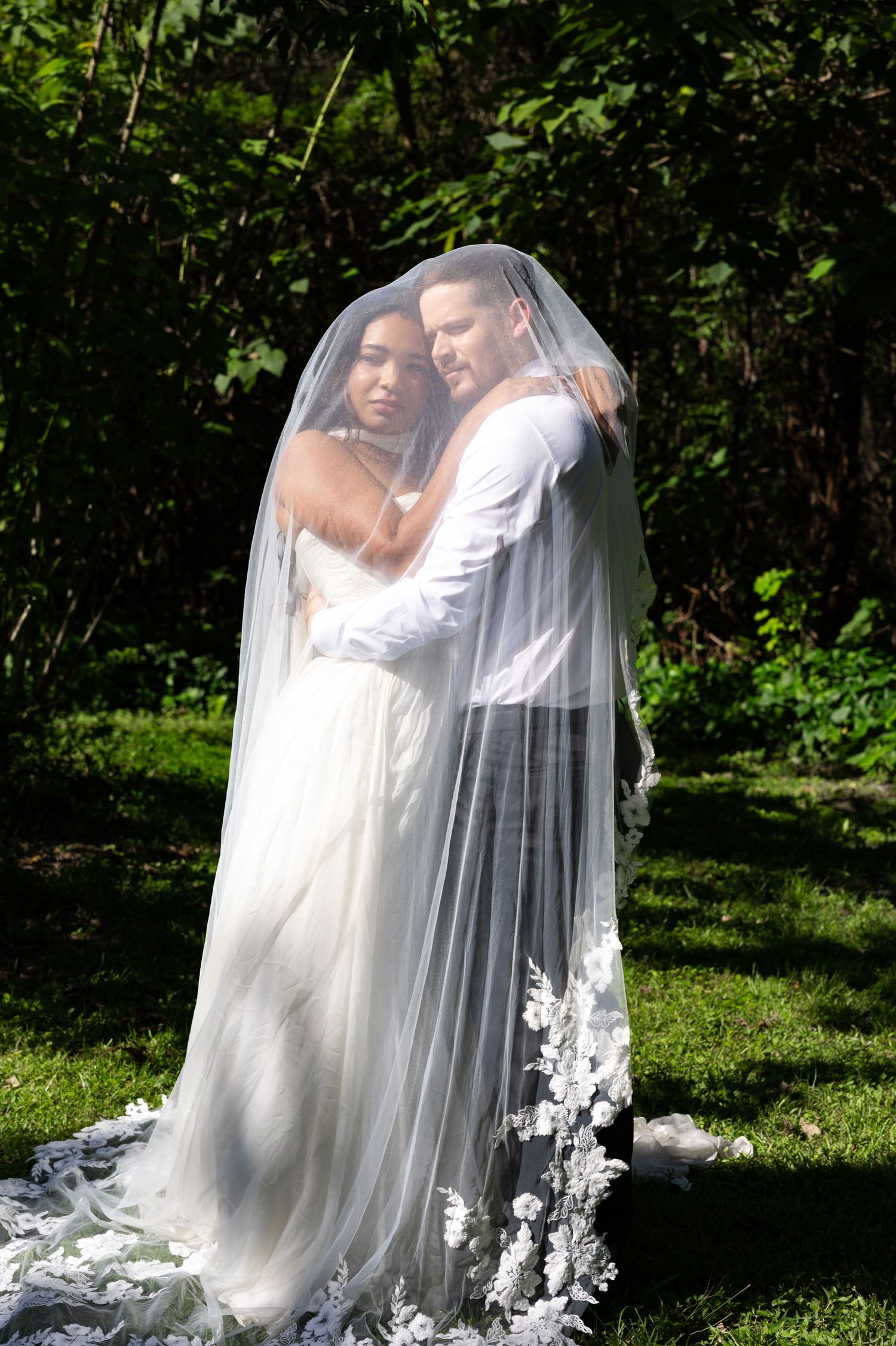 Bride and groom share romantic embrace under flowing white lace veil in lush green garden wedding photo.