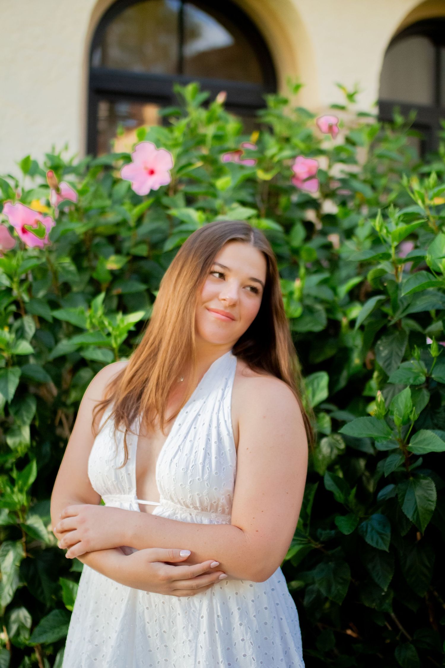 Young woman in white halter dress poses outdoors near lush green hibiscus bushes with pink blooms.