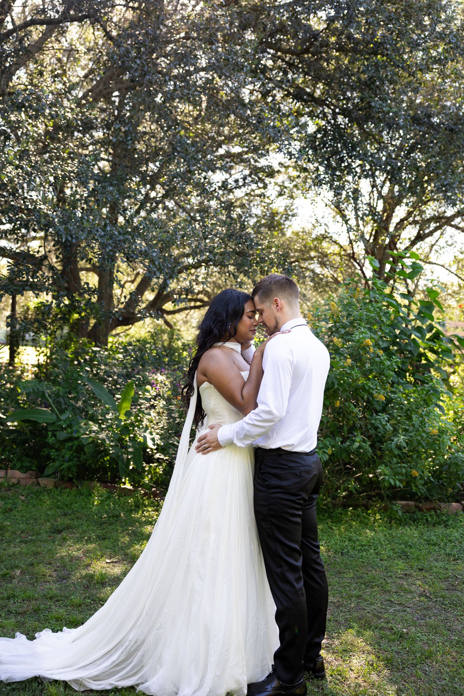 Bride and groom embrace in lush garden setting, bride wearing elegant white gown with long train.