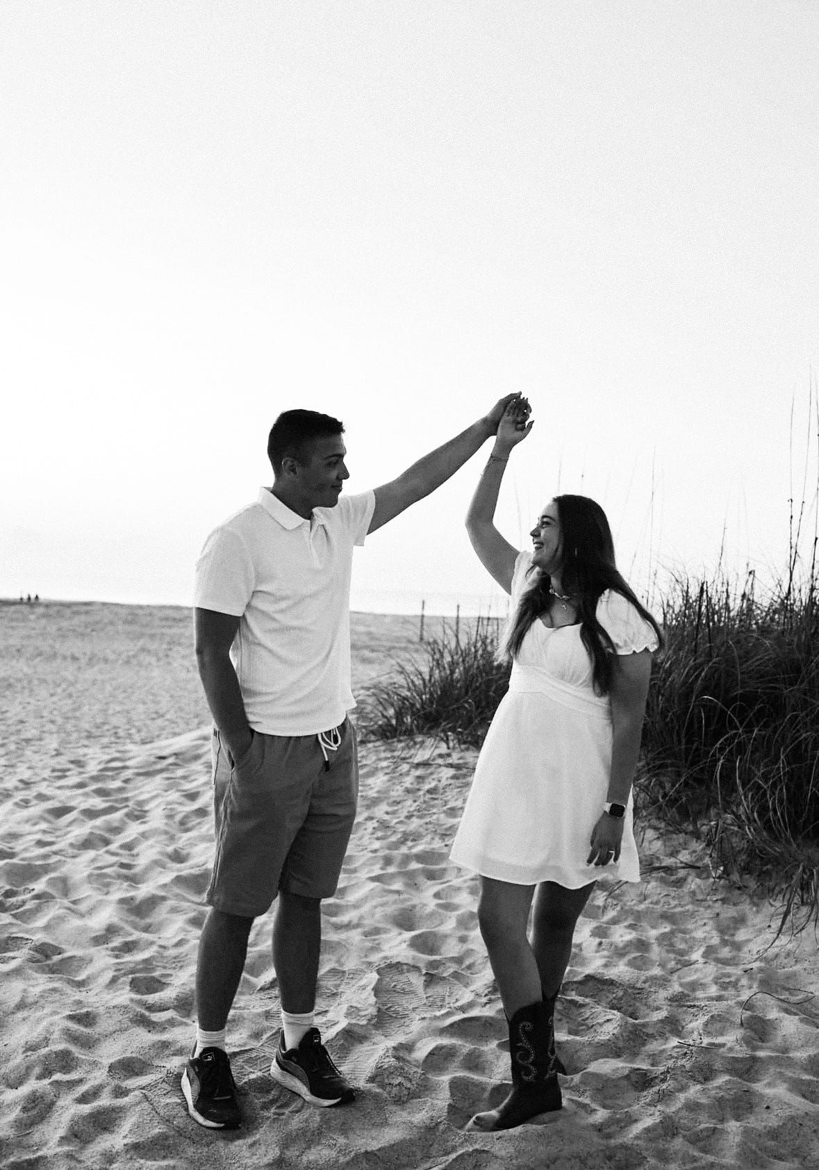 Black and white photo of a couple dancing on a sandy beach, man spinning woman in a white dress near dunes.