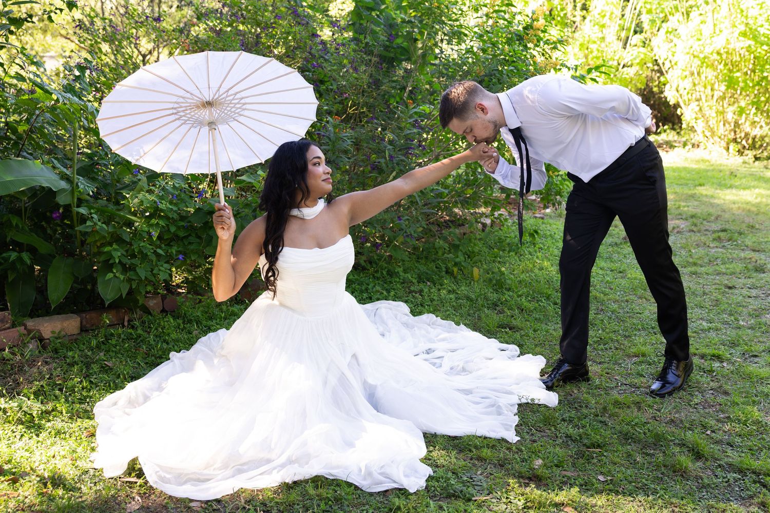 Bride in white gown holds parasol while groom kisses her hand outdoors in lush green garden setting.
