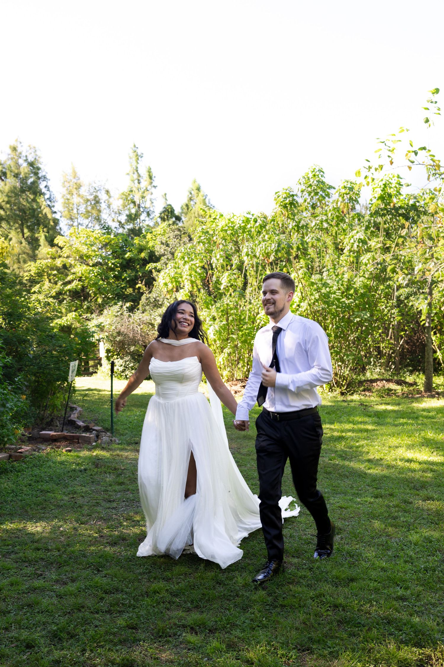 Happy interracial newlywed couple holding hands and walking in lush green garden, bride in white gown, groom in black suit.