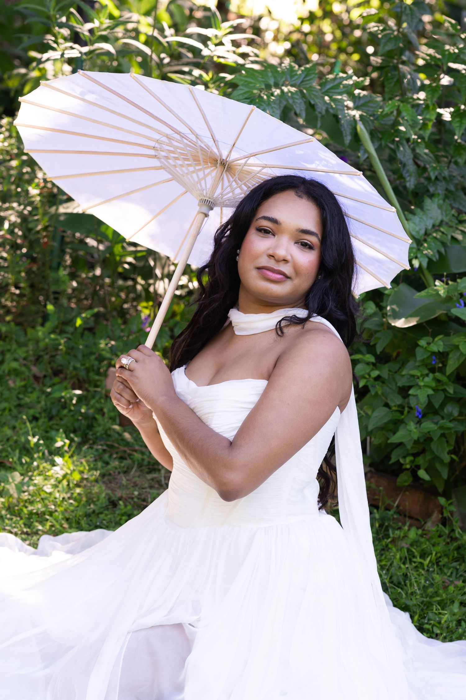 A bride in a white dress holds a white parasol umbrella while posing outdoors in a lush green garden.
