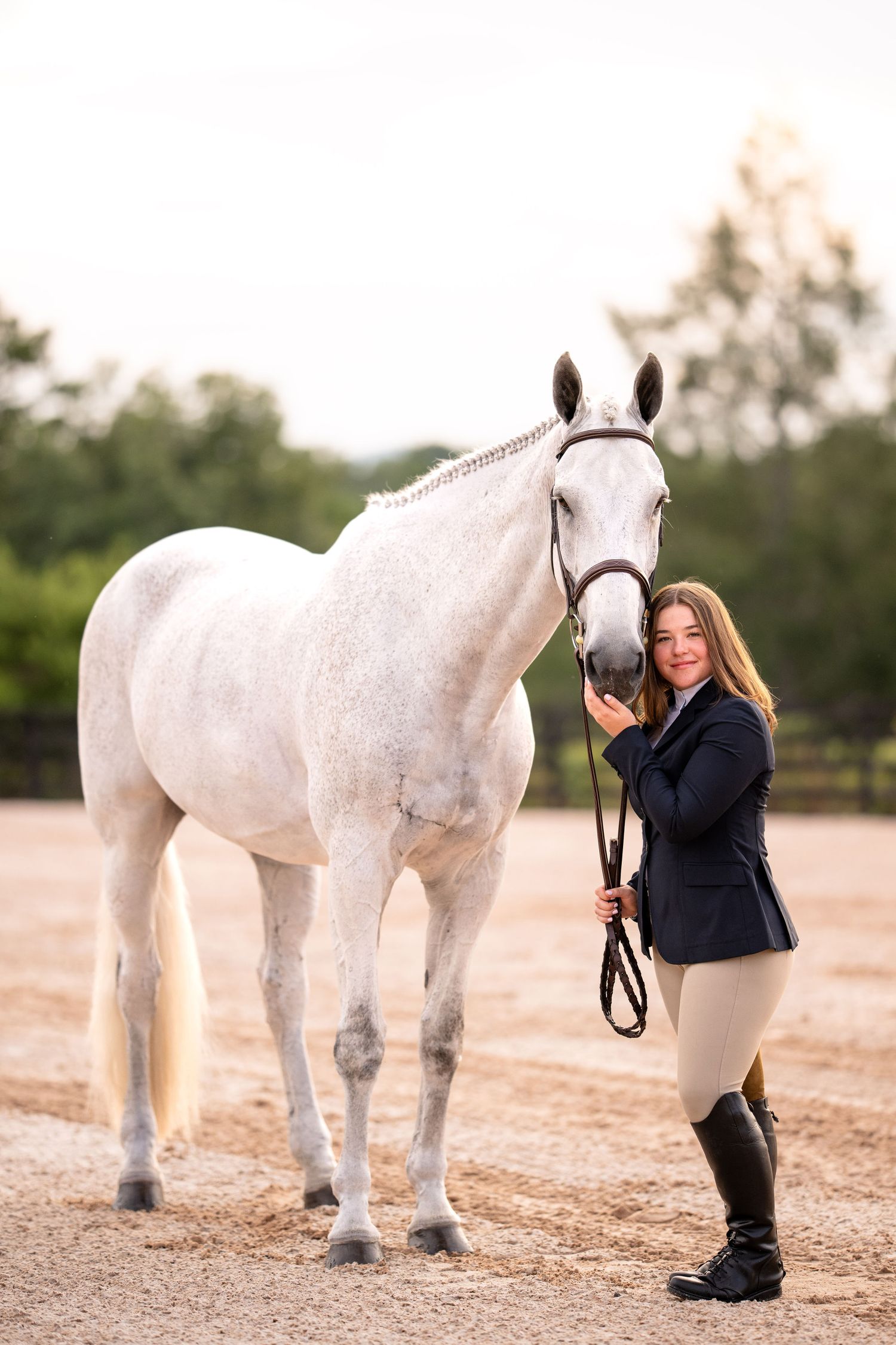 Young woman in equestrian attire standing beside a white horse on a sandy riding arena.