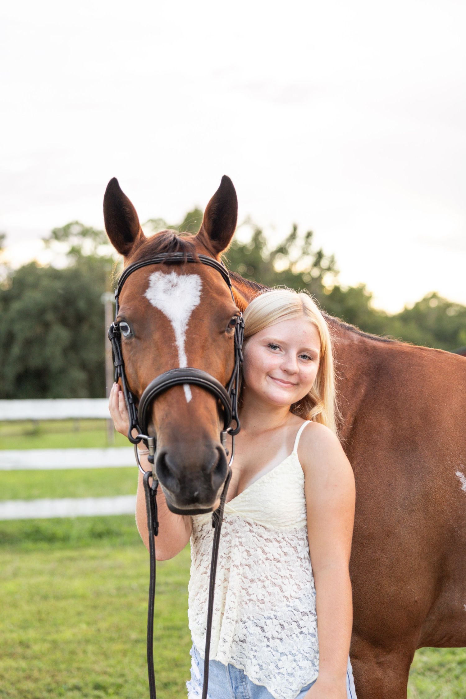 Smiling blonde girl posing with a chestnut horse with a heart-shaped blaze in a green pasture.
