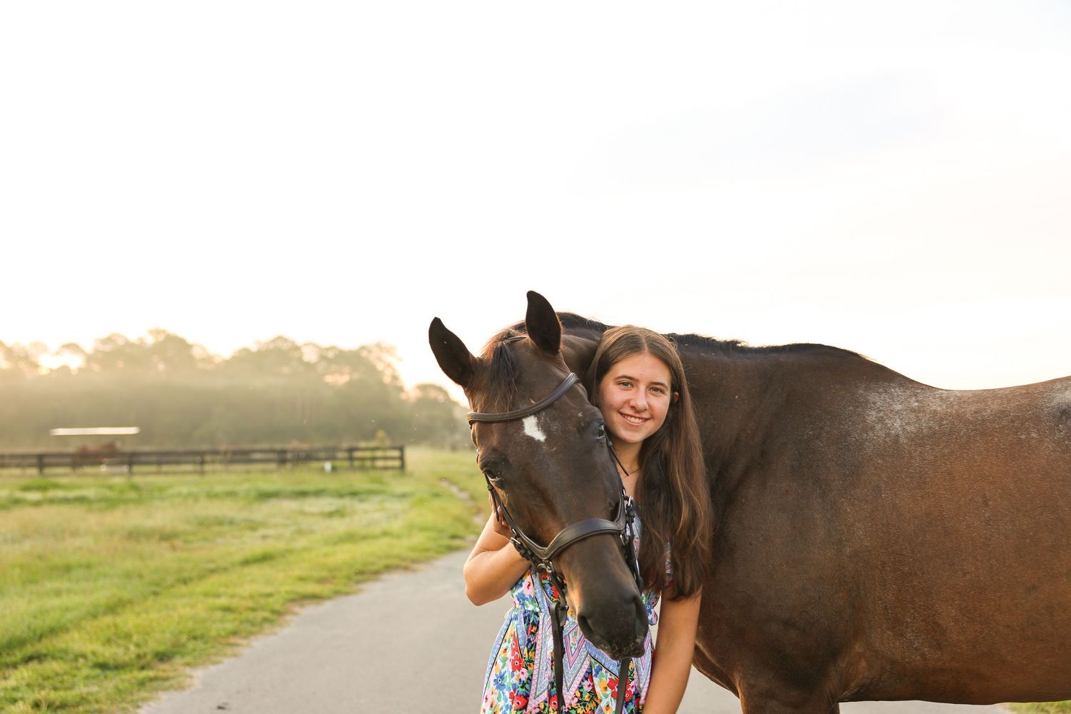 Young girl smiling beside a dark brown horse on a sunny farm path with green fields in the background.