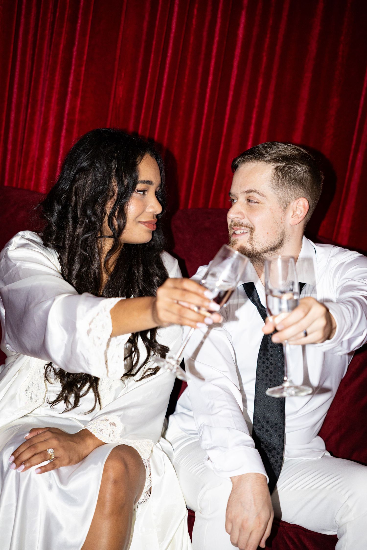 Couple in white wedding attire toasting champagne glasses, smiling at each other against red curtain backdrop.