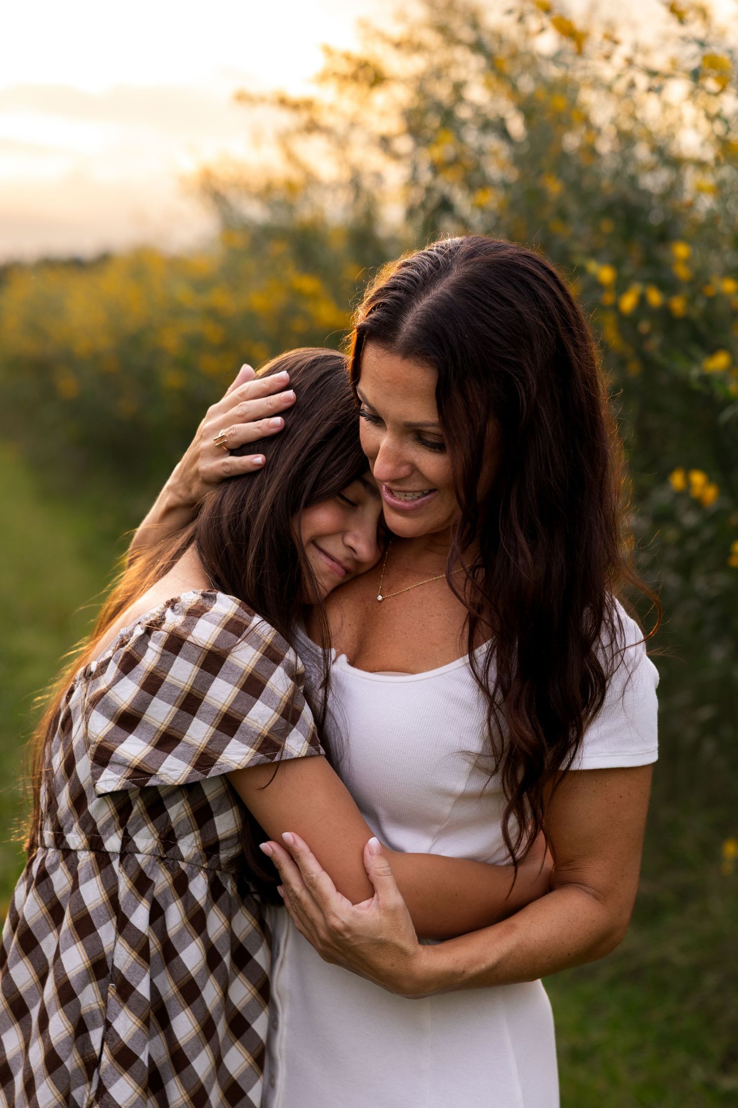 A mother and daughter share a warm embrace in a golden field at sunset, wearing white and plaid dresses.