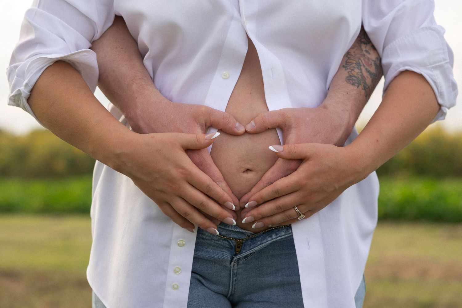 Couple forming heart shape with hands on pregnant belly outdoors in white shirt and jeans.
