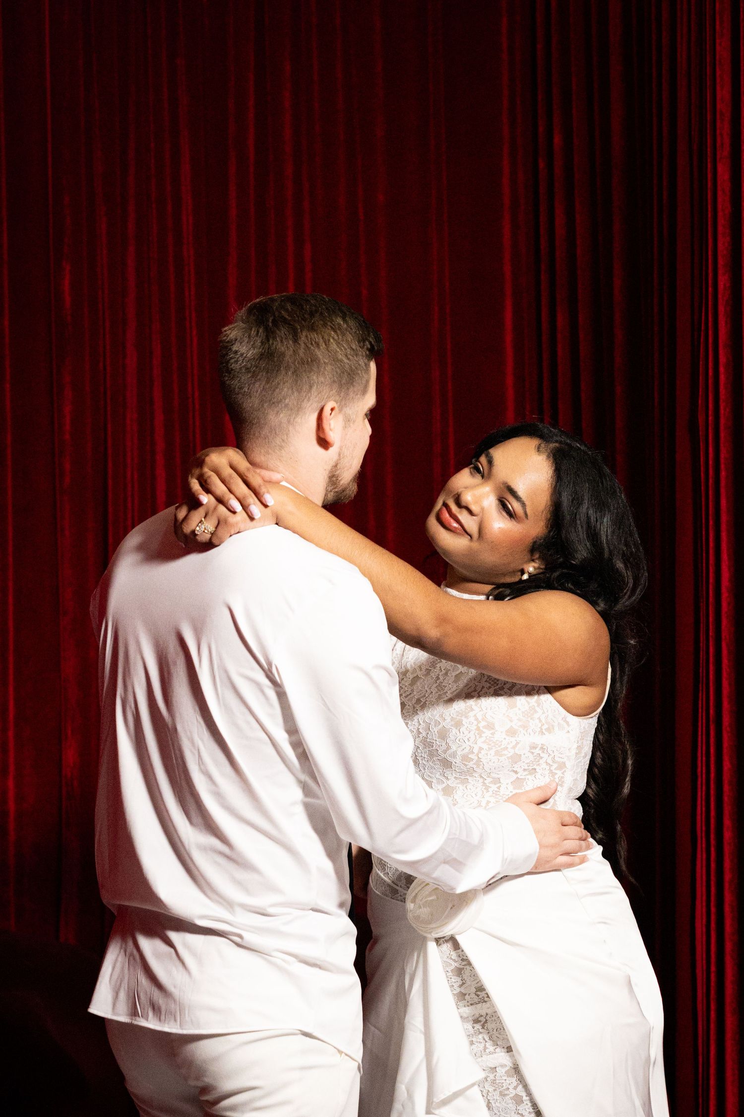 Couple in white attire embracing before red curtain backdrop, woman smiling warmly at partner.
