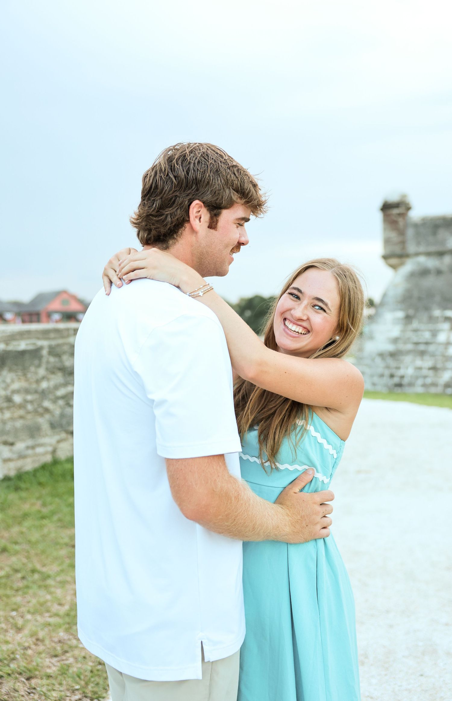 A smiling couple embraces outdoors near a historic stone wall, woman in turquoise dress, man in white shirt.