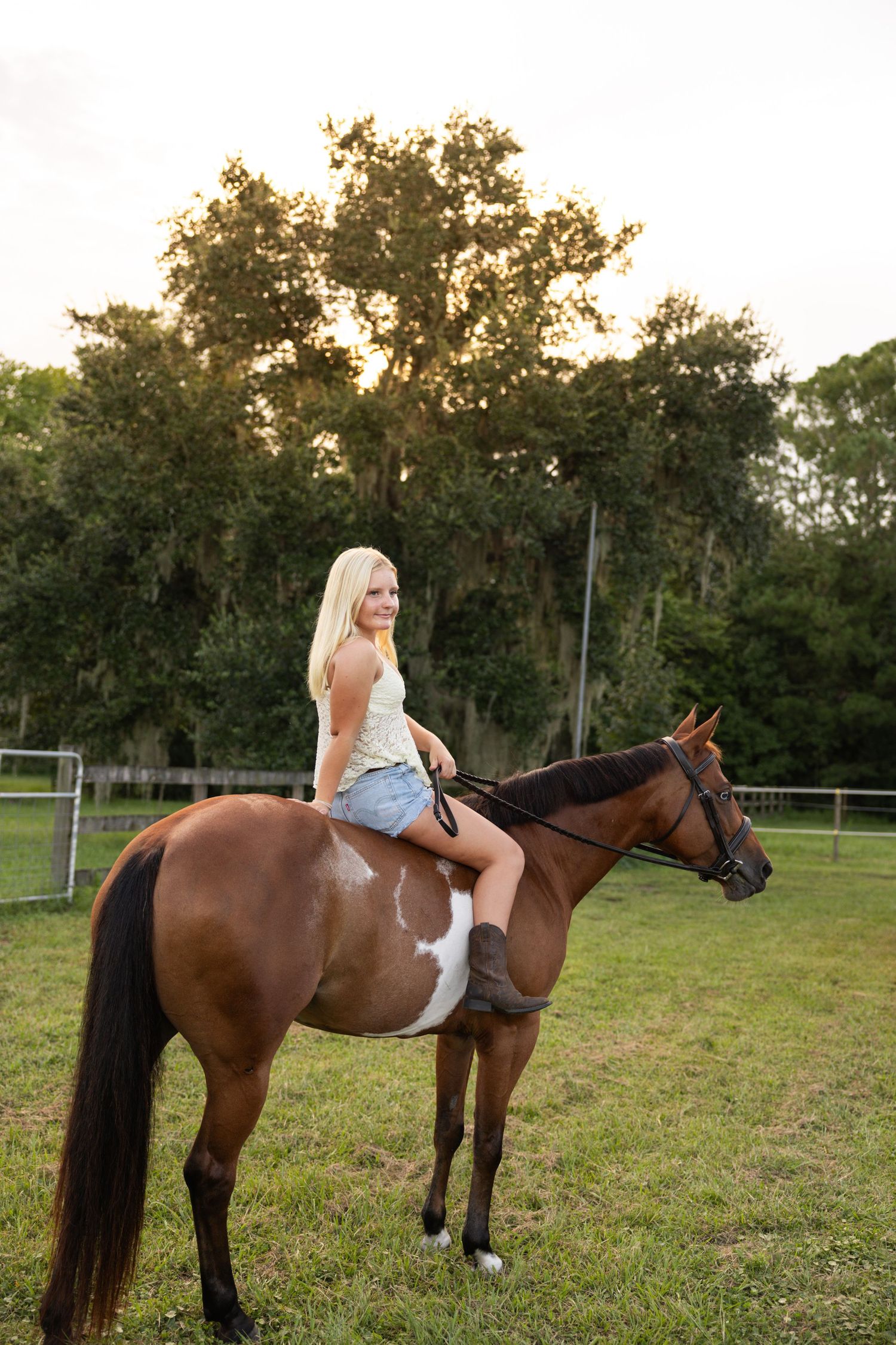 Blonde woman riding a brown and white paint horse in a green pasture with trees in the background.
