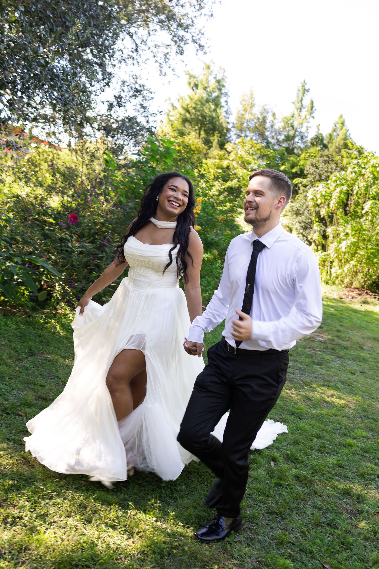 Happy interracial couple in wedding attire holding hands and running through a lush green garden on their wedding day.