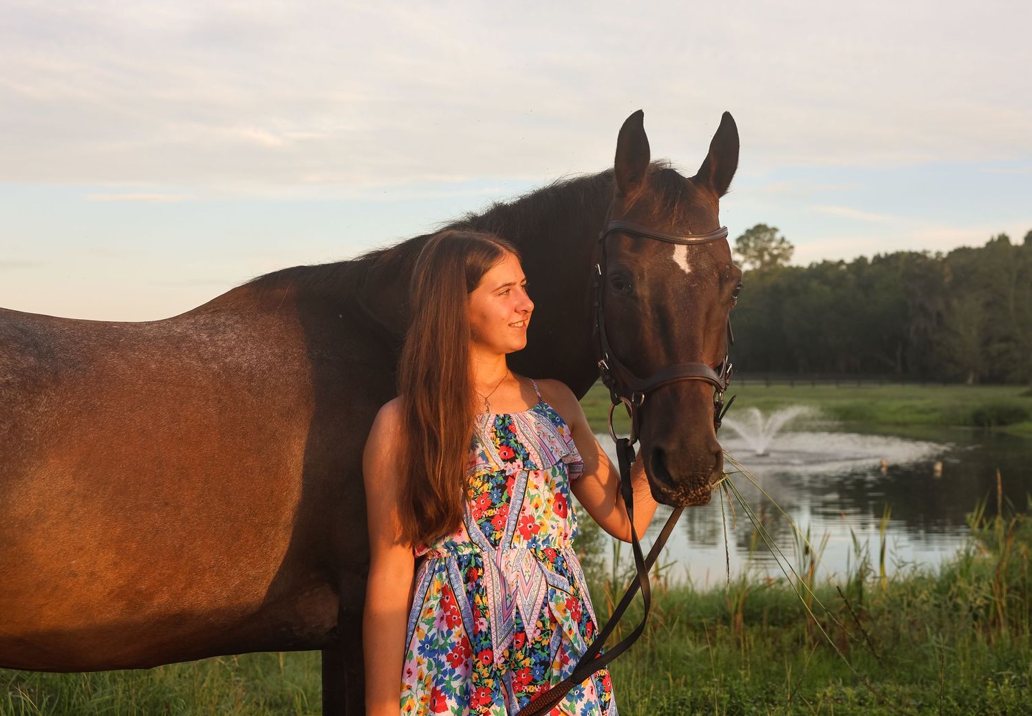 Young woman in floral dress standing with a dark bay horse in a sunny green field near a pond at golden hour.