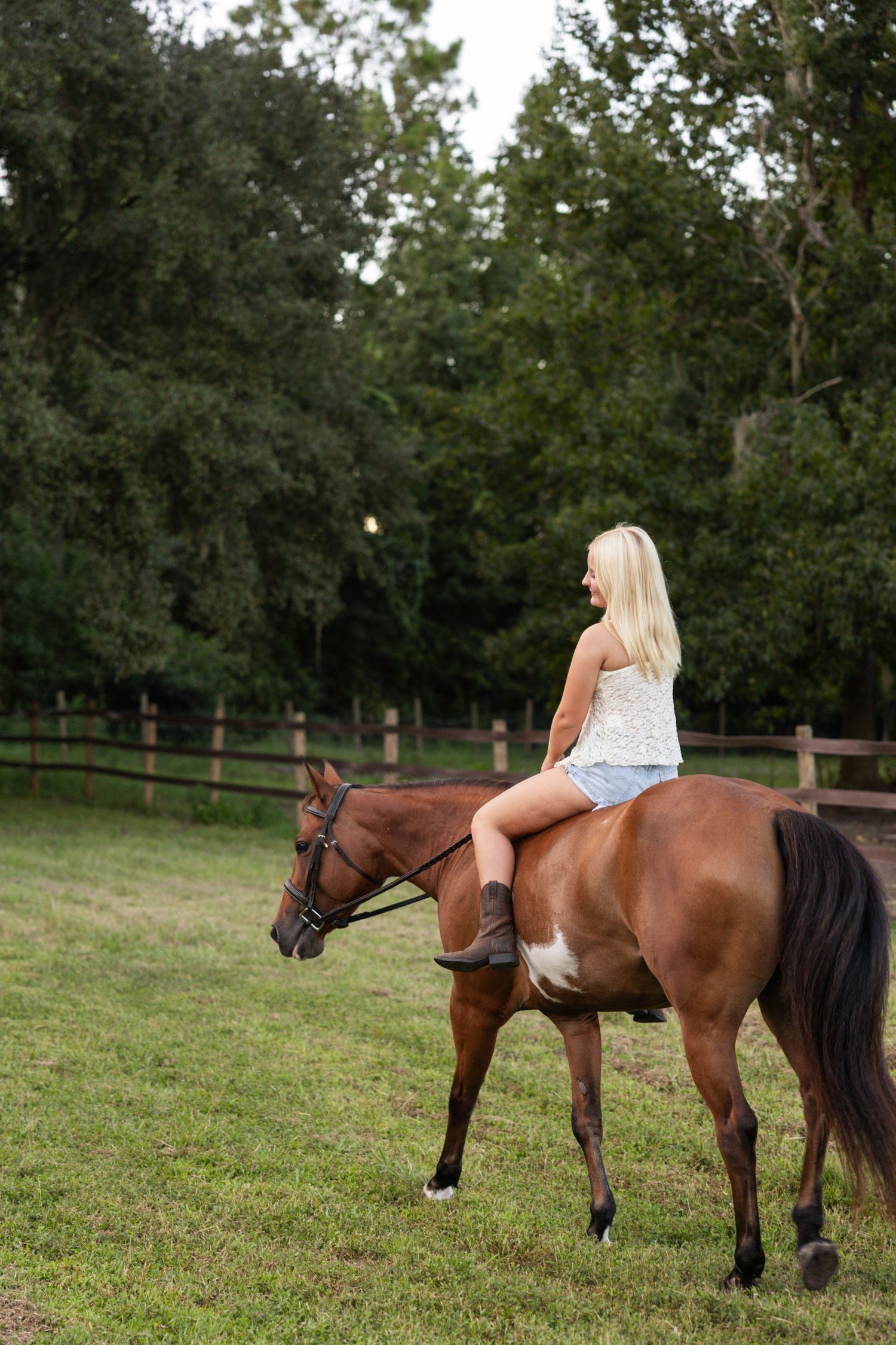 Blonde woman in denim shorts riding a bay horse in a green paddock with wooden fence and trees.