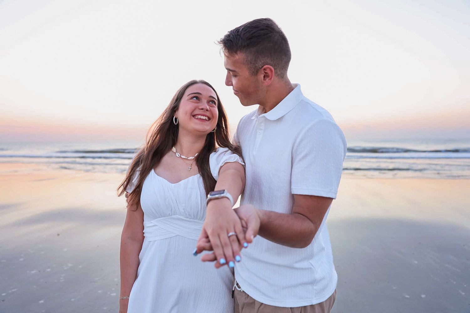 Happy couple embracing on a beach at sunset, woman in white dress showing engagement ring.