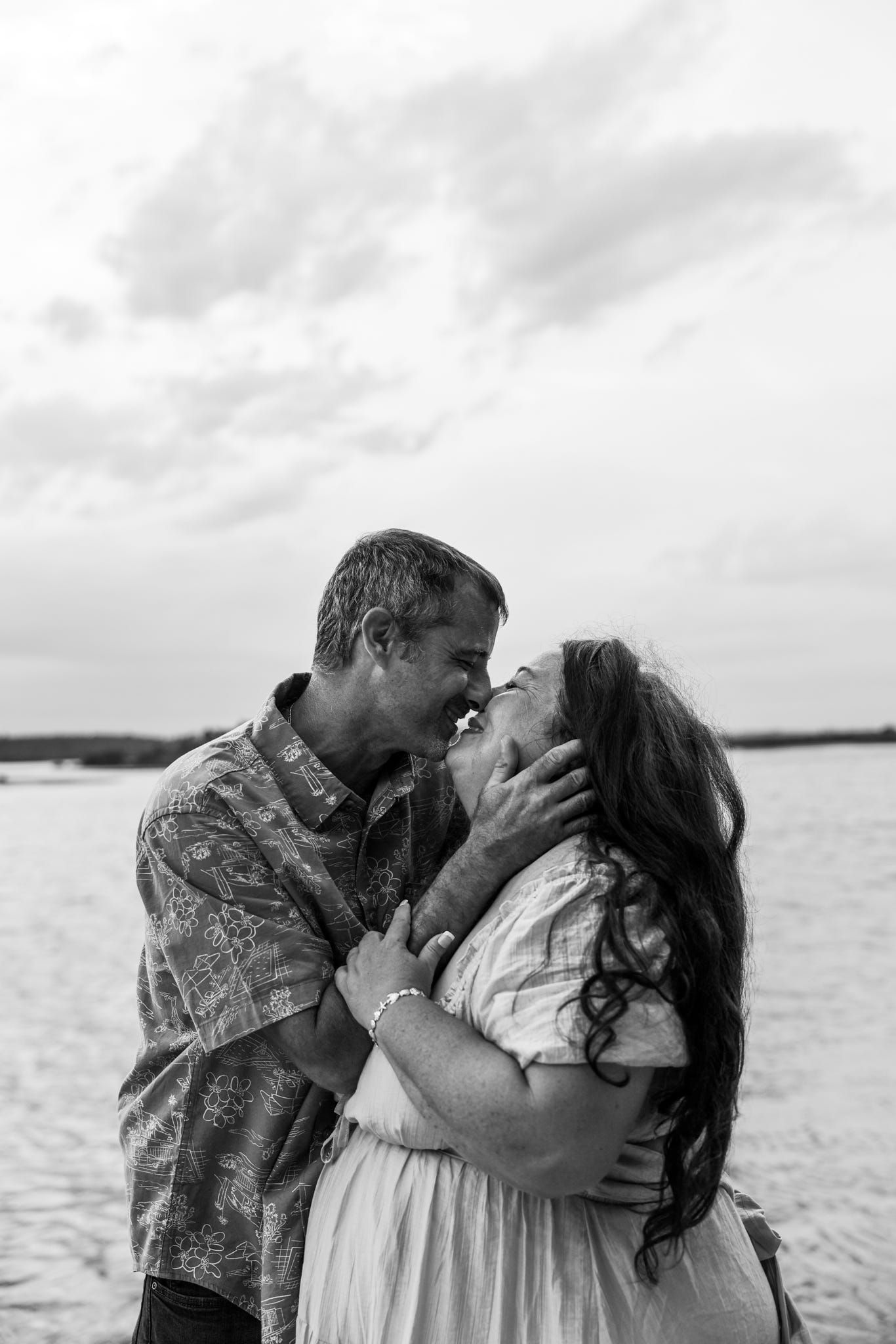 Black and white photo of a couple sharing a romantic kiss on a beach with cloudy skies in the background.