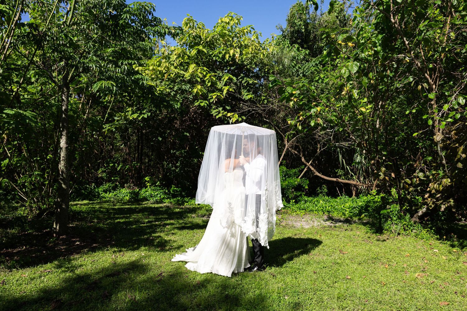 Bride in white wedding dress and veil standing in lush tropical garden on sunny day.