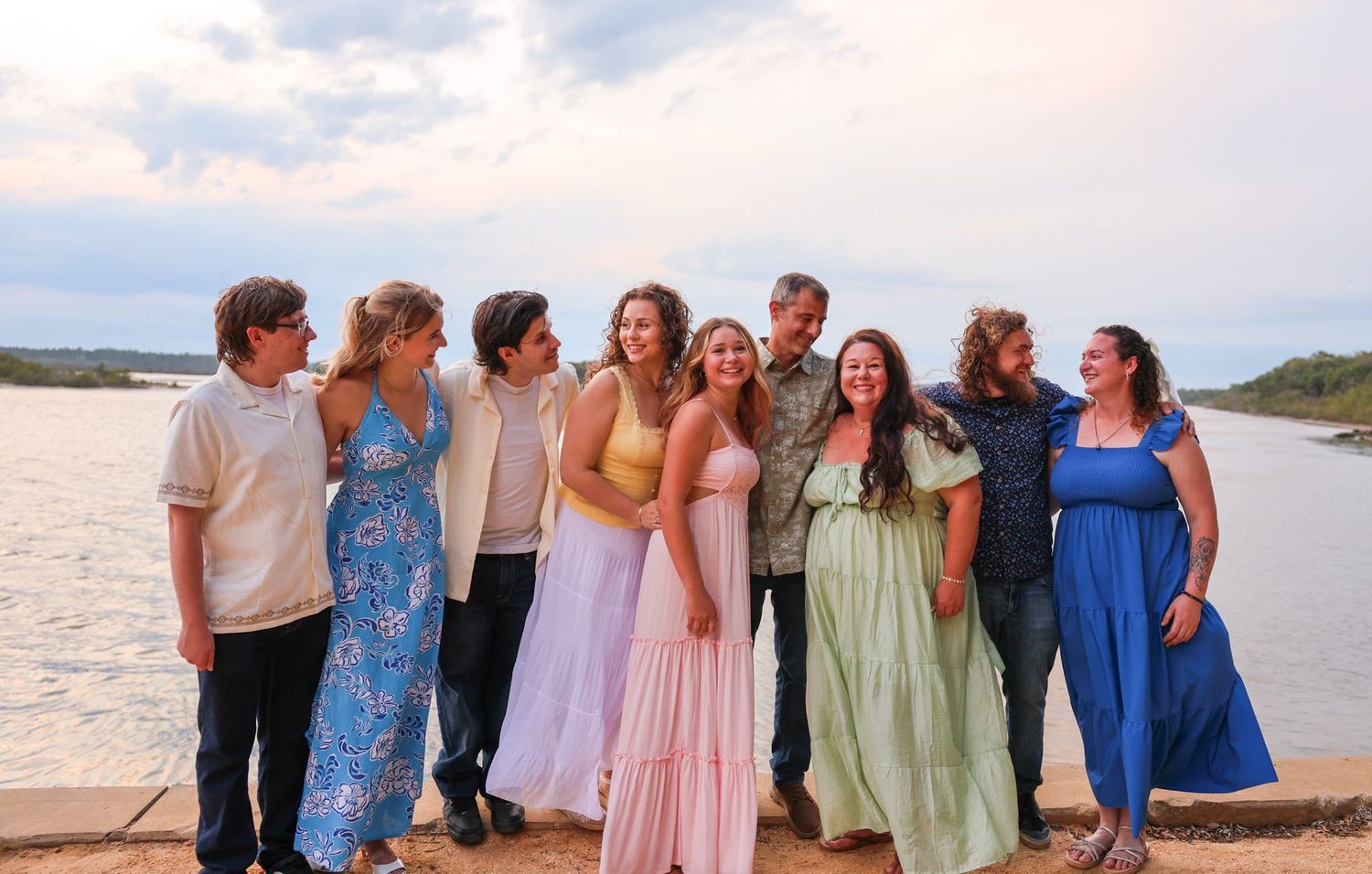 Group of nine people smiling together outdoors near a lake with a cloudy sky backdrop.