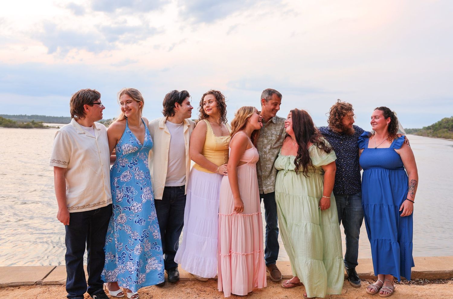 Large family group posing together outdoors near a lake or river, wearing colorful summer dresses and casual attire.