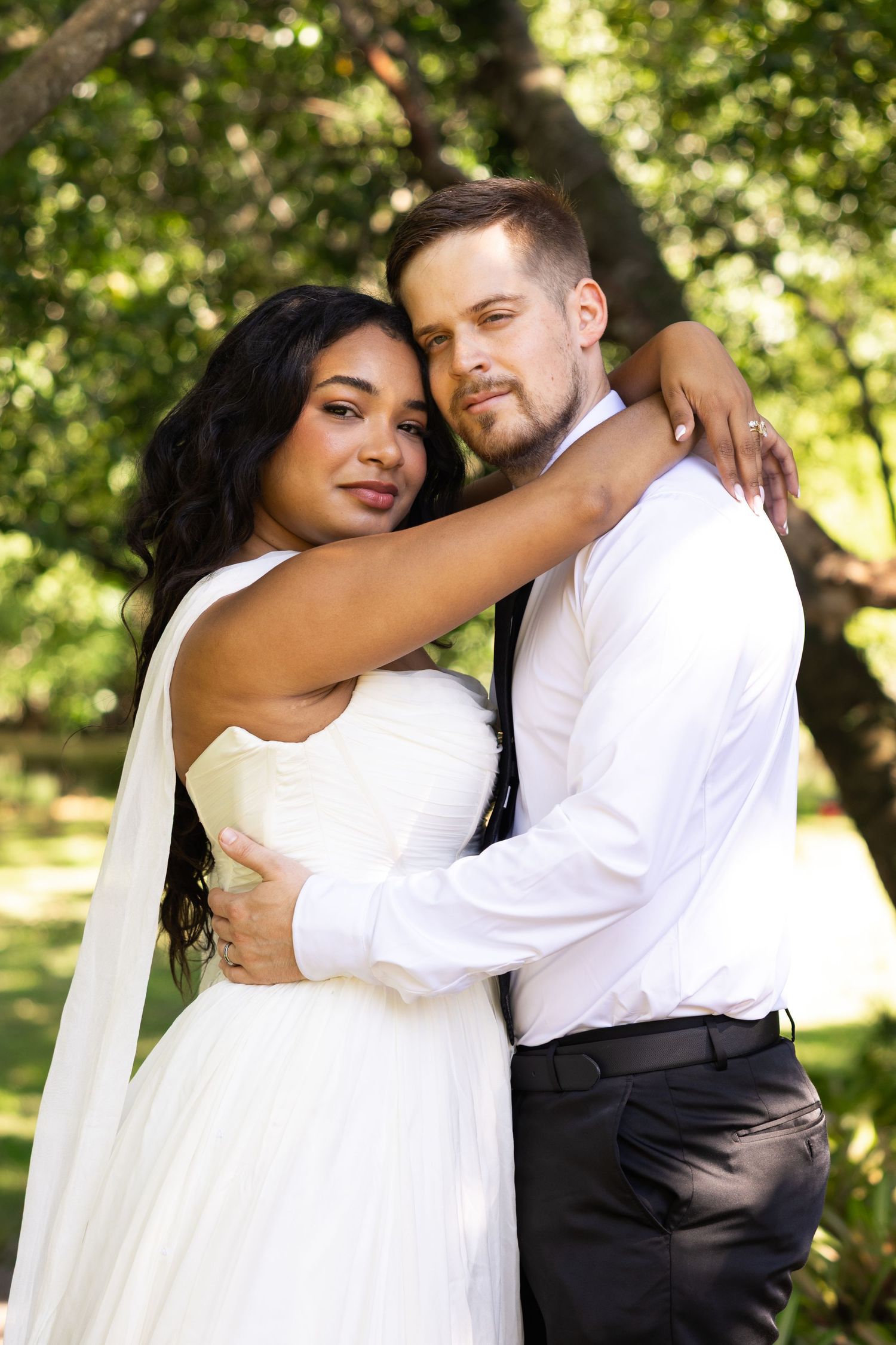 A bride and groom embrace outdoors, she in a white gown, he in a white shirt and black pants, surrounded by lush greenery.