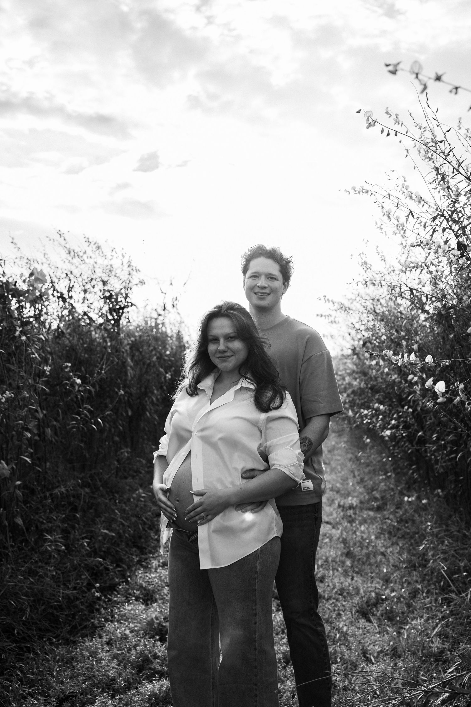 Black and white maternity photo of a couple outdoors, woman cradling baby bump, man standing behind her smiling.