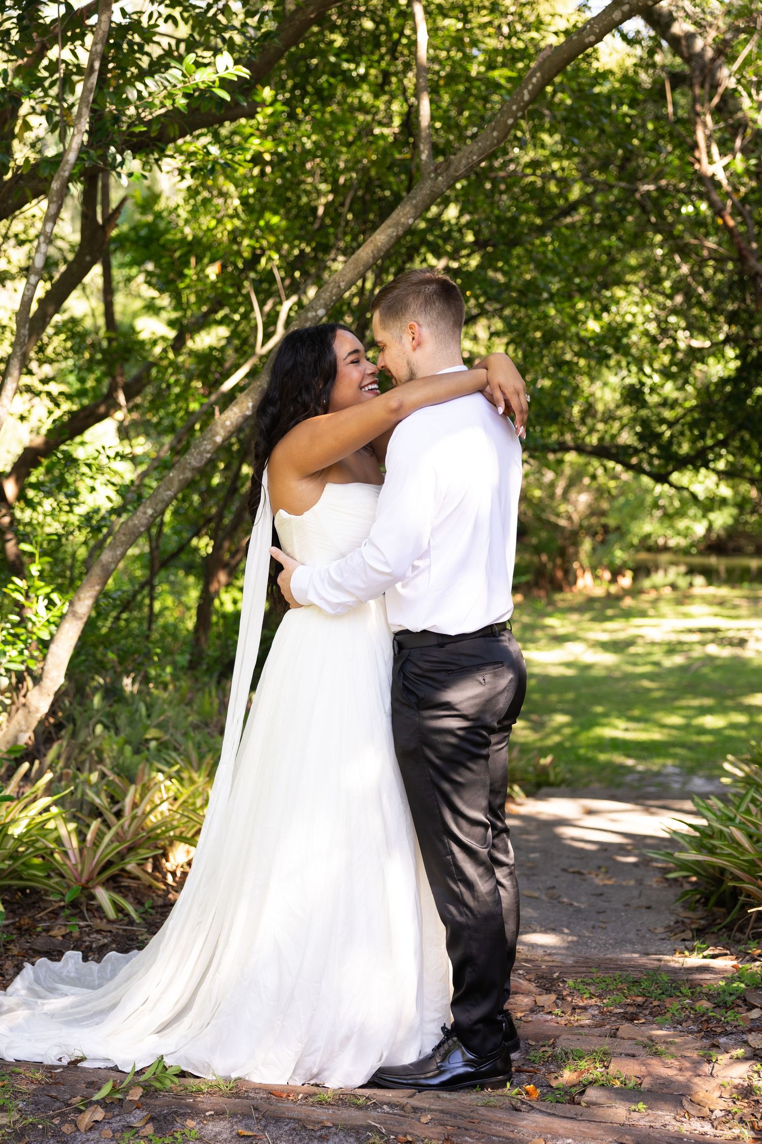 A bride in a white gown embraces her groom in a lush green garden, smiling joyfully on their wedding day.