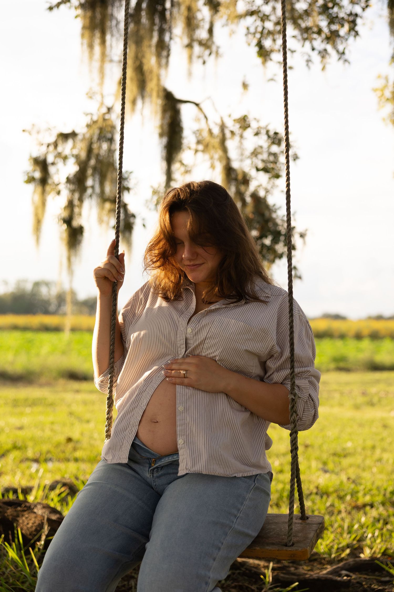 Pregnant woman sitting on a rope swing outdoors, cradling her baby bump, wearing a white shirt and jeans at golden hour.