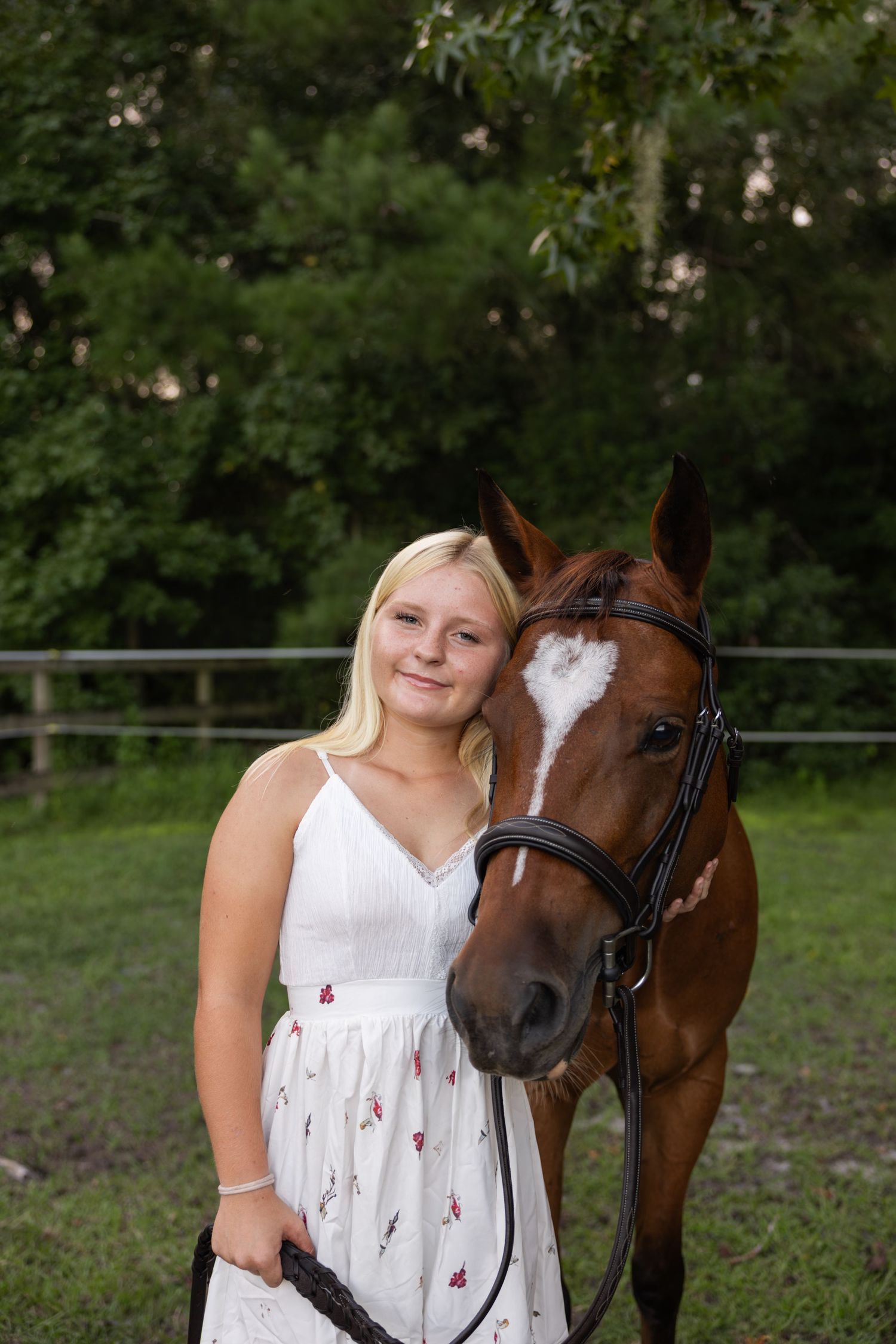Blonde girl in white floral dress posing with chestnut horse wearing bridle in green pasture.