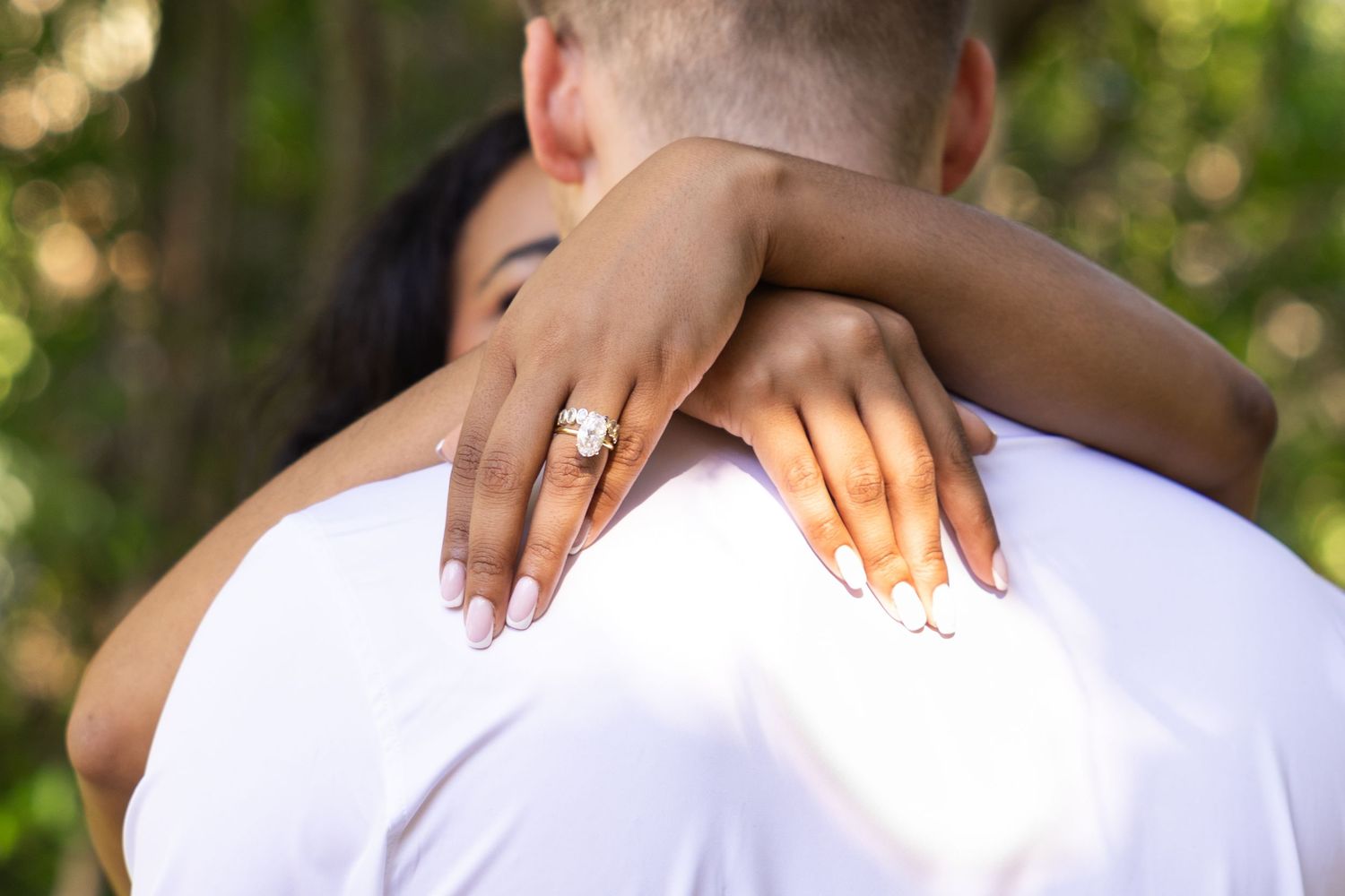 Couple embracing outdoors, woman displaying large diamond engagement ring with white manicured nails on man's shoulder.