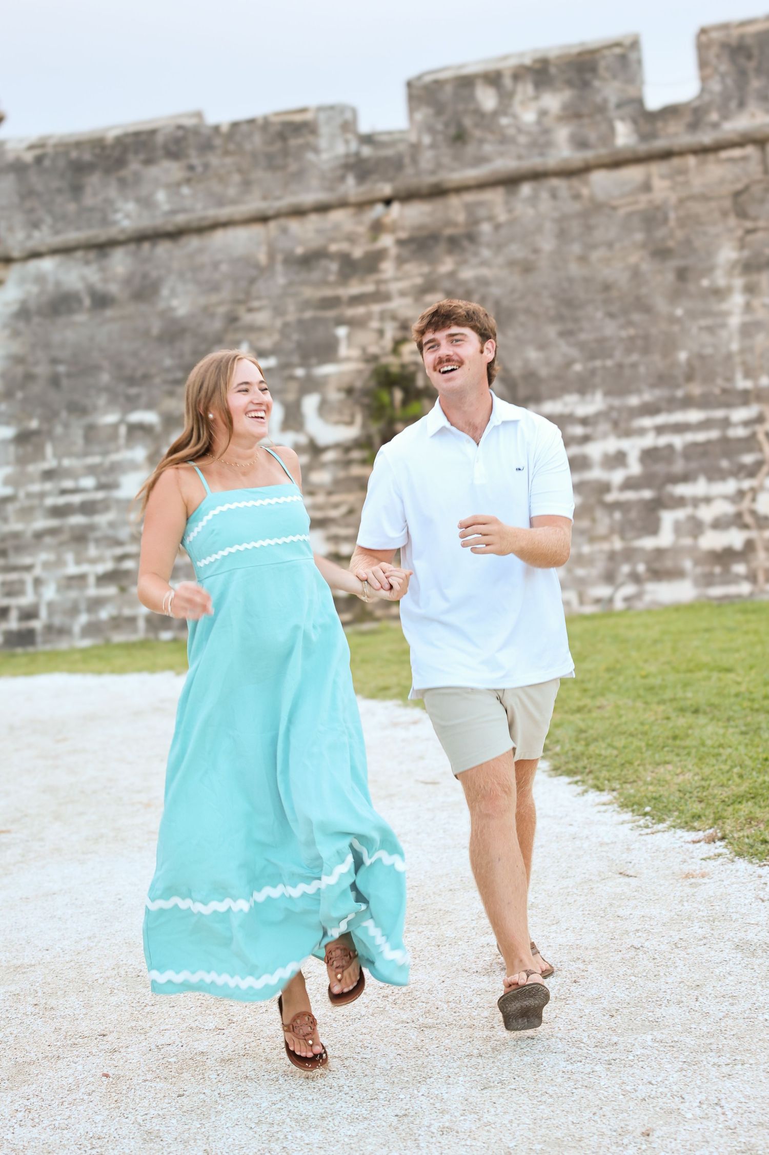 A smiling couple holds hands and walks near a stone wall, woman in teal maxi dress, man in white polo and khaki shorts.