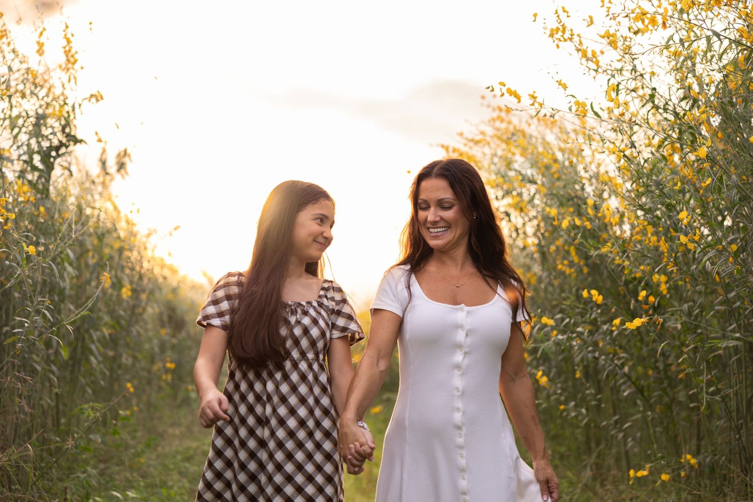 Two women walking hand in hand through a sunlit field of yellow wildflowers, smiling at each other.
