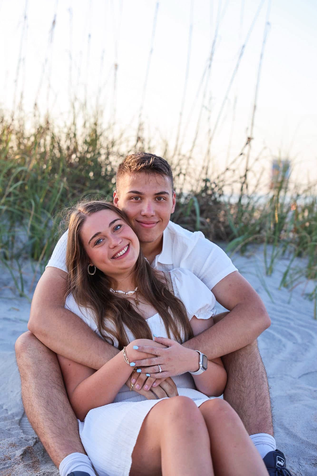 Young couple sitting together on sandy beach dunes, smiling and embracing, wearing white outfits at sunset.