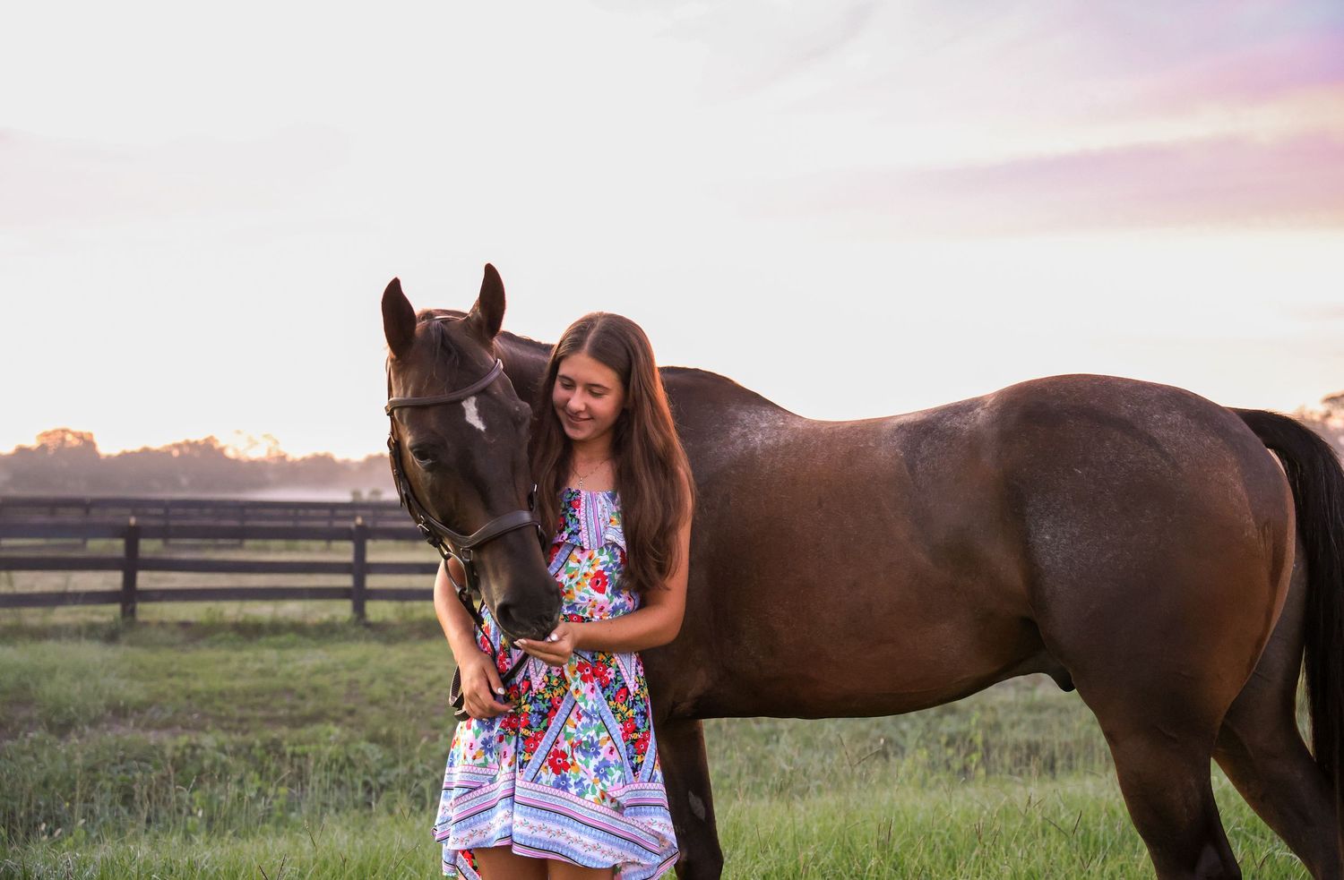 Young girl in floral dress stands beside a dark brown horse in a green pasture at sunset.