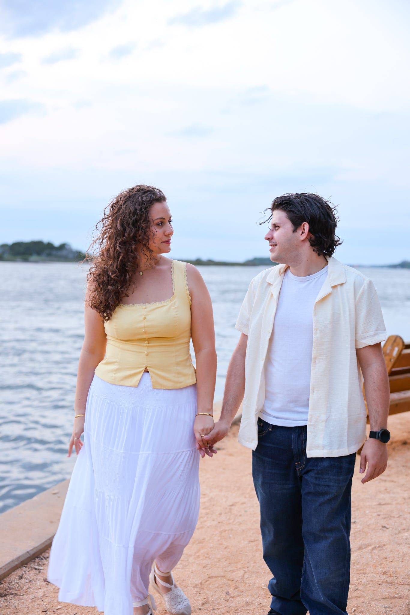 A couple holds hands and walks along a waterfront dock at dusk, smiling at each other.