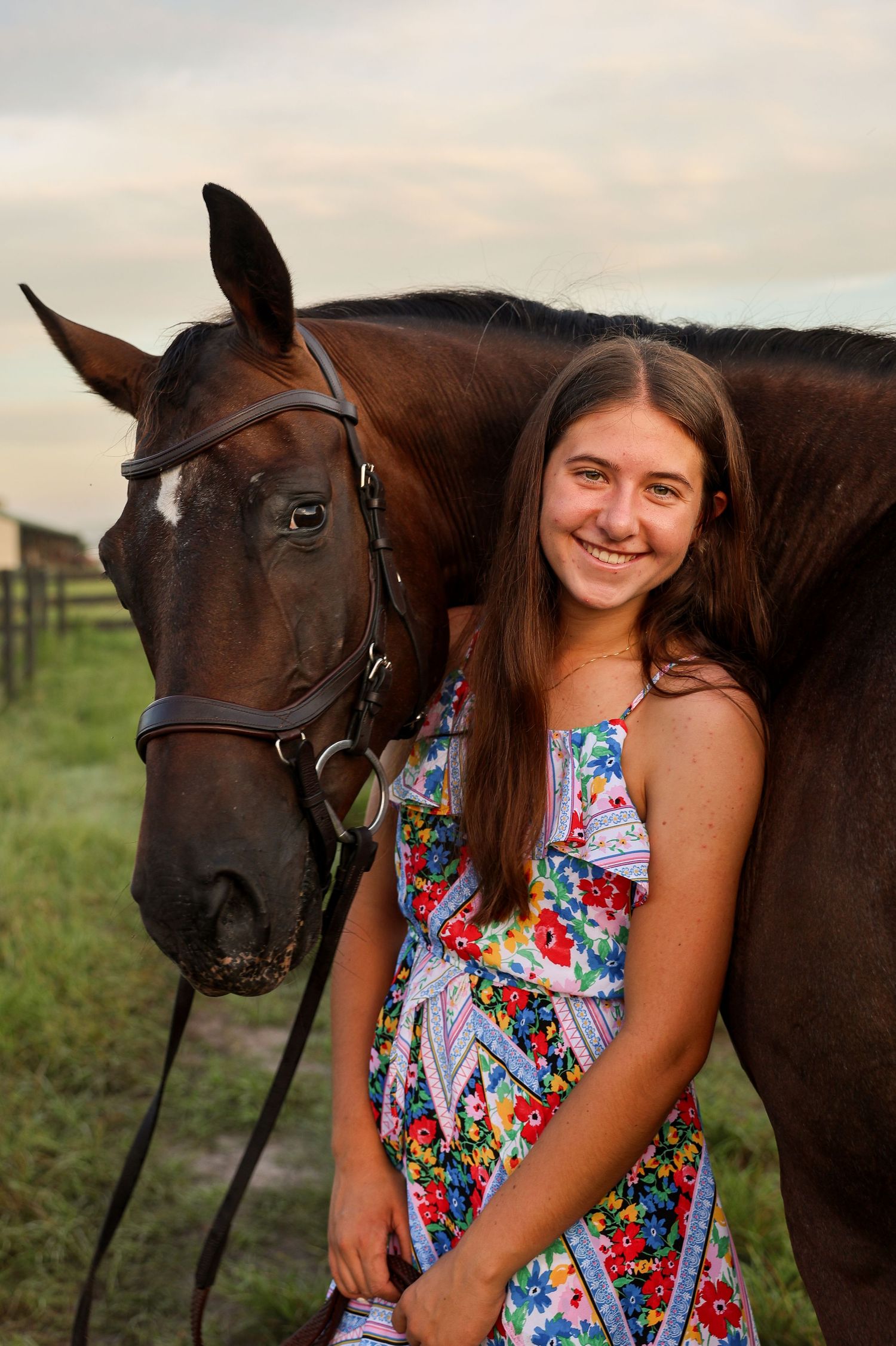 Smiling teenage girl in a floral dress posing with a dark brown horse wearing a bridle on a farm.