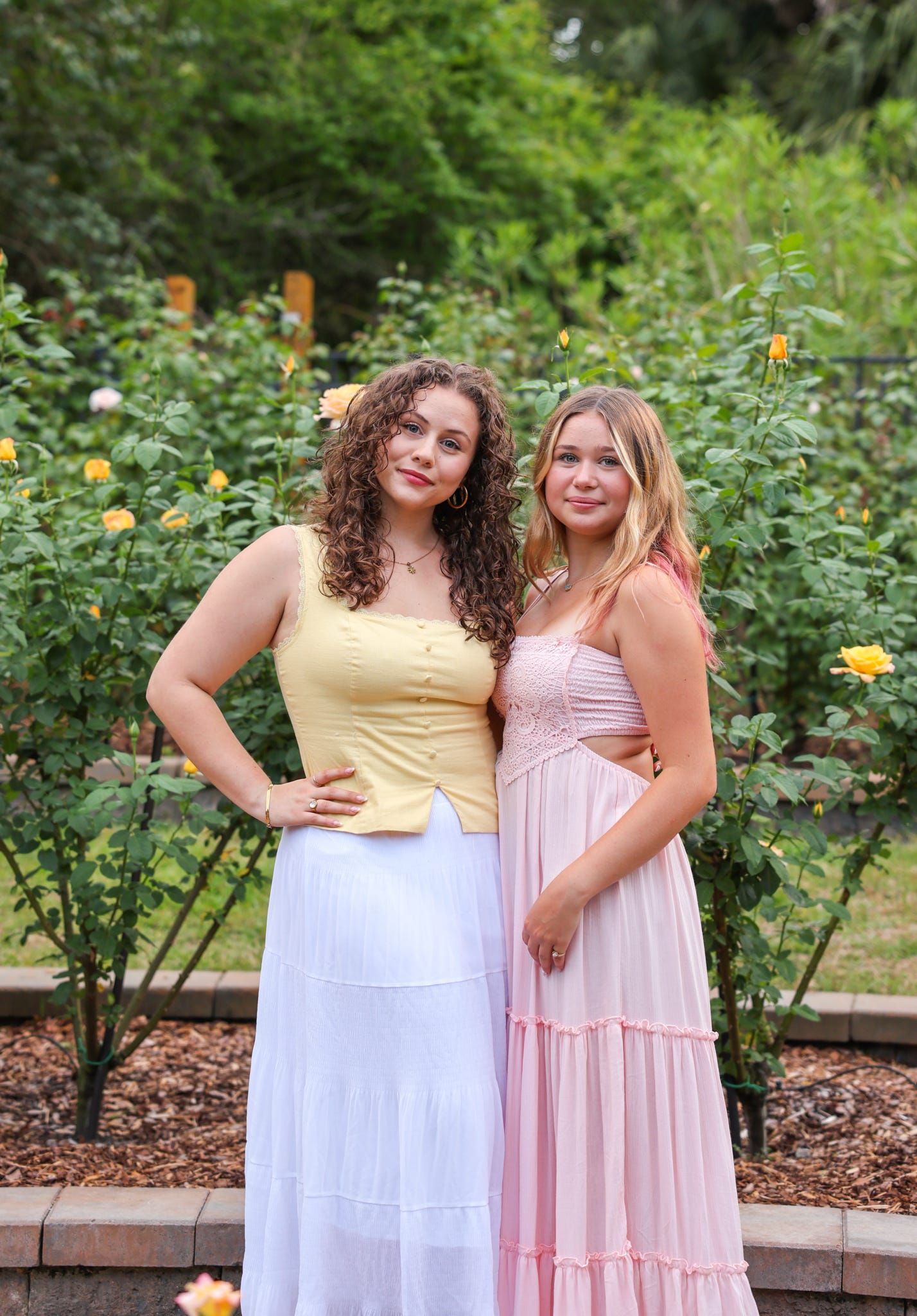 Two young women in elegant dresses pose together in a garden with yellow roses blooming in the background.