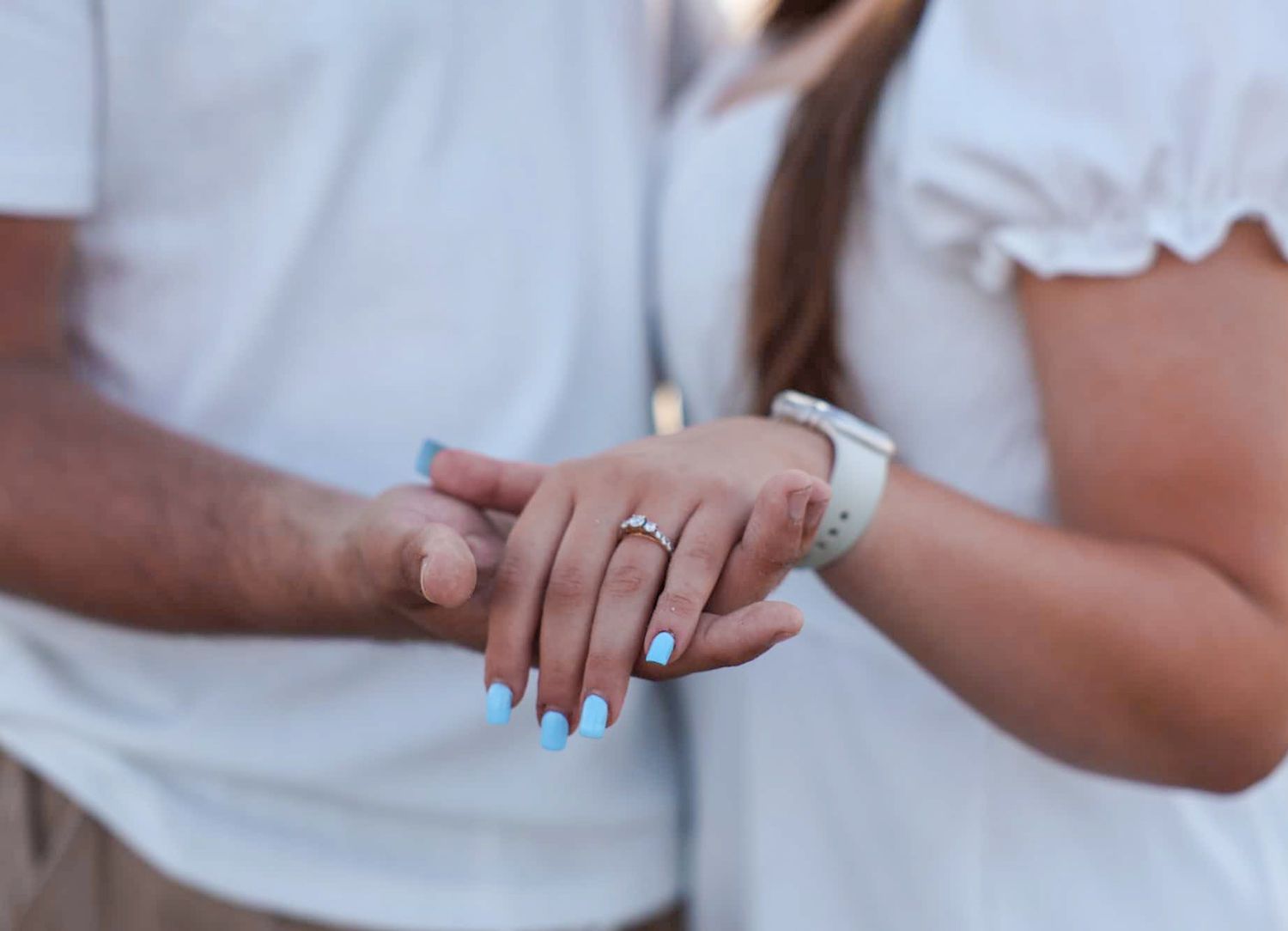Couple holding hands with blue manicure and diamond engagement ring, both wearing white outfits.