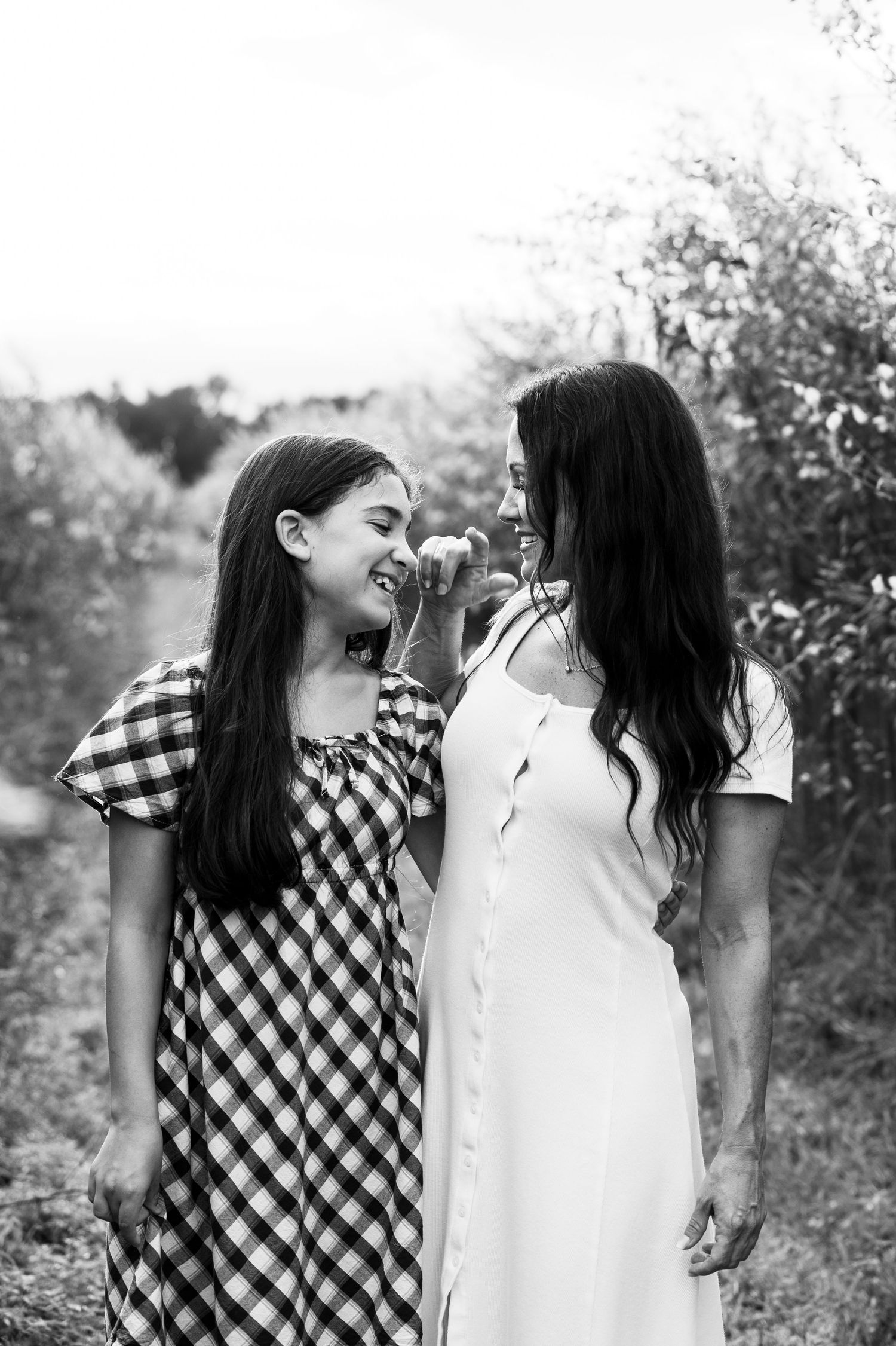 Black and white photo of two young women smiling at each other outdoors, wearing dresses, surrounded by trees.