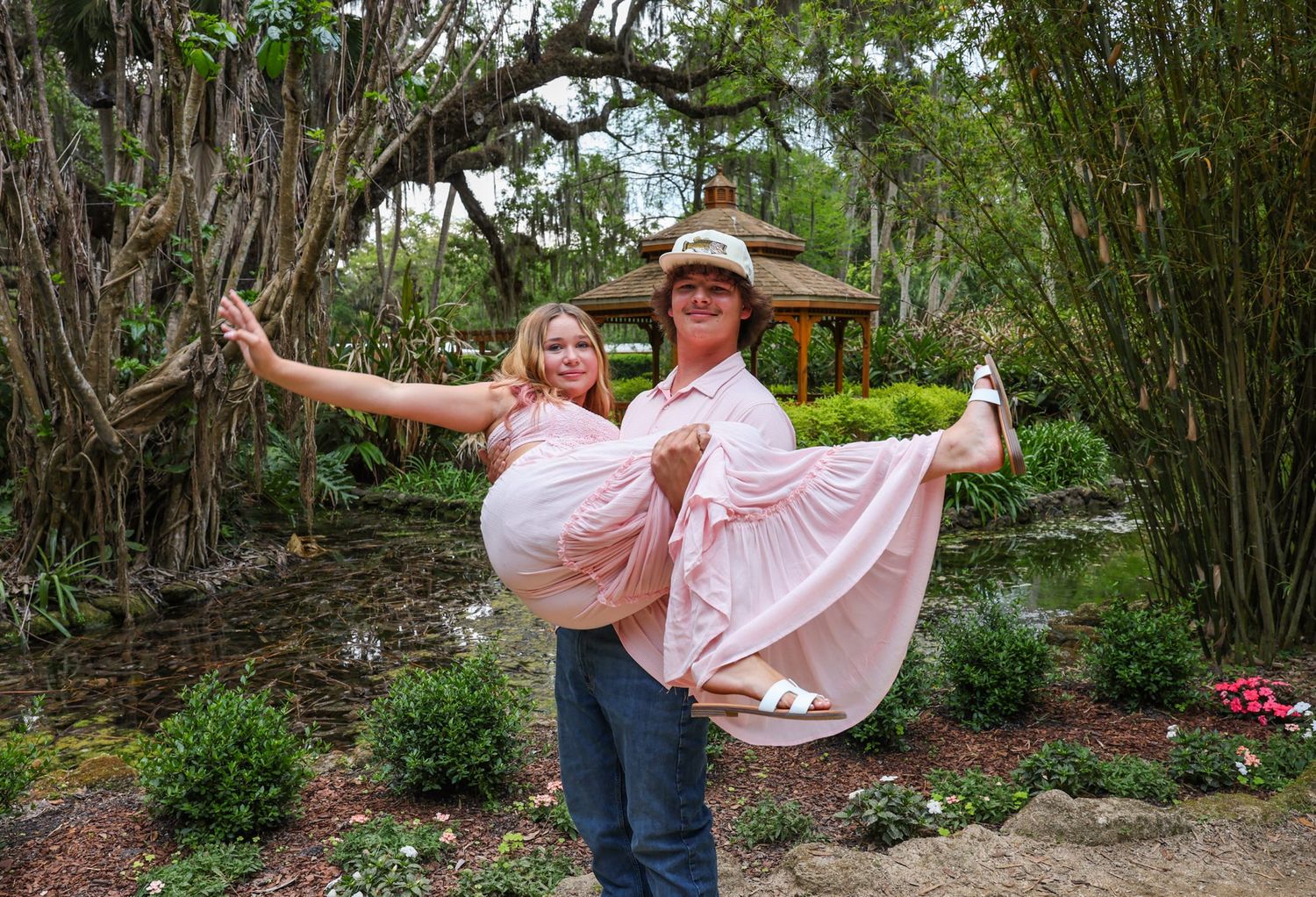 Couple posing in lush garden, man lifting woman in pink dress under sprawling trees near gazebo.