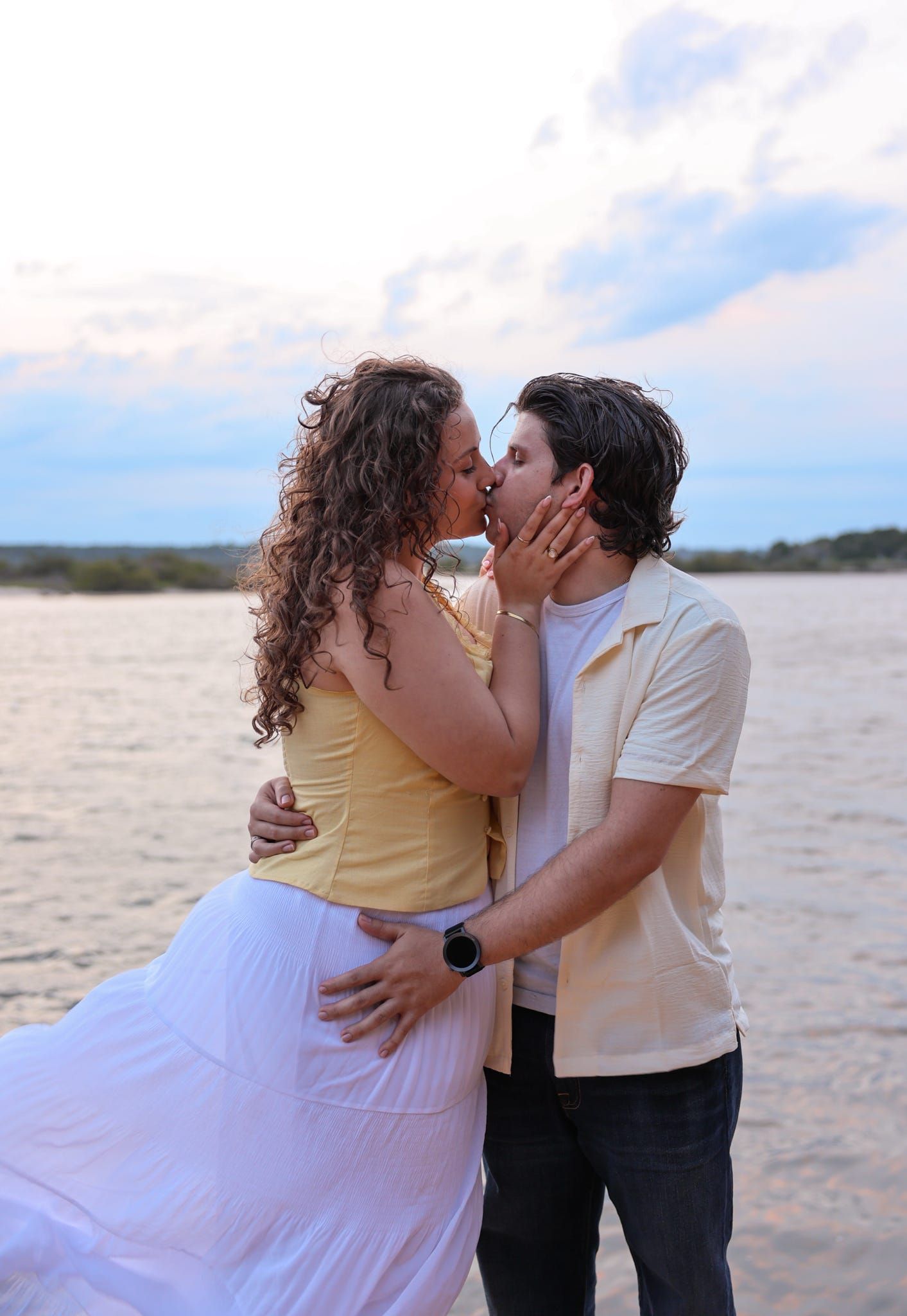 Couple sharing a romantic kiss by the water at sunset, woman in yellow top and white skirt, man in cream shirt.