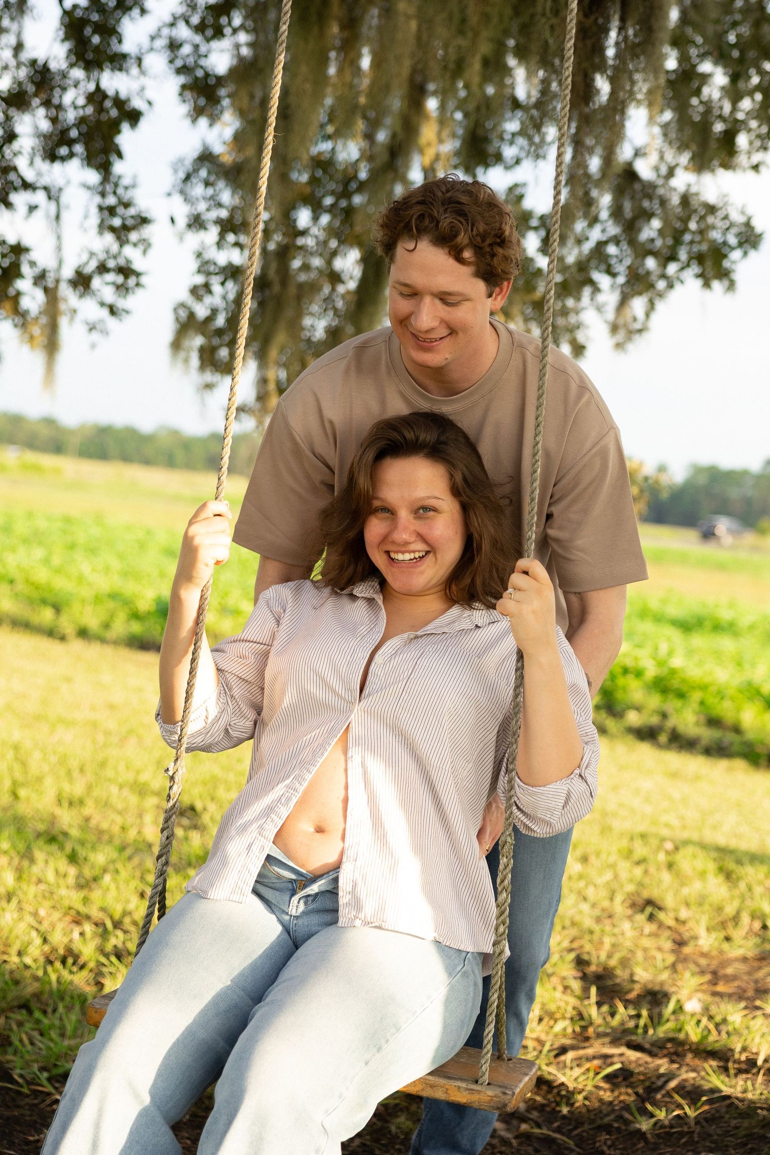 Pregnant woman smiling on rope swing outdoors while partner stands behind her in sunny green field.