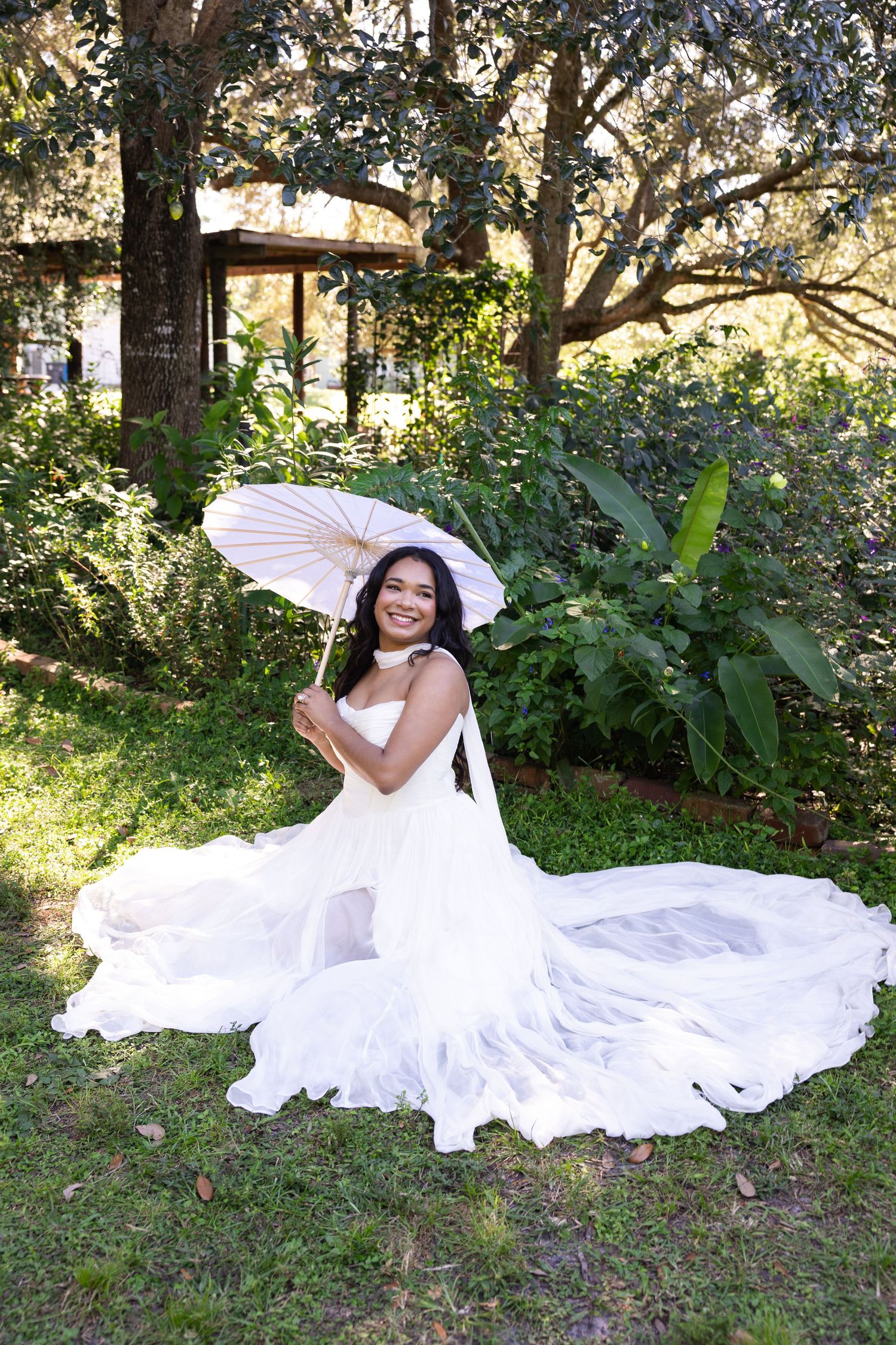 Smiling bride in white gown holding parasol umbrella seated on grass in lush garden setting.