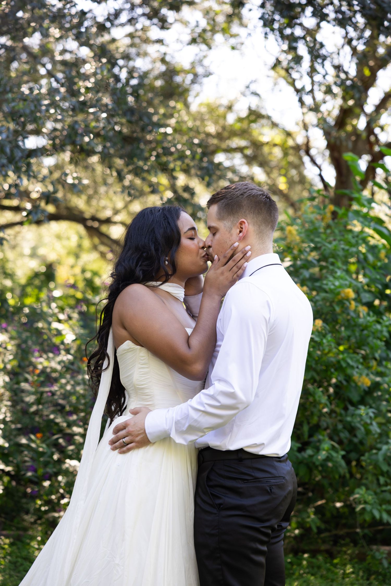 Interracial couple sharing a romantic kiss in a lush garden, bride in white gown and groom in white shirt.