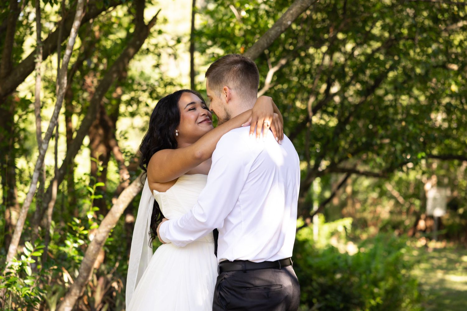 A bride and groom share a romantic embrace outdoors surrounded by lush green trees on their wedding day.
