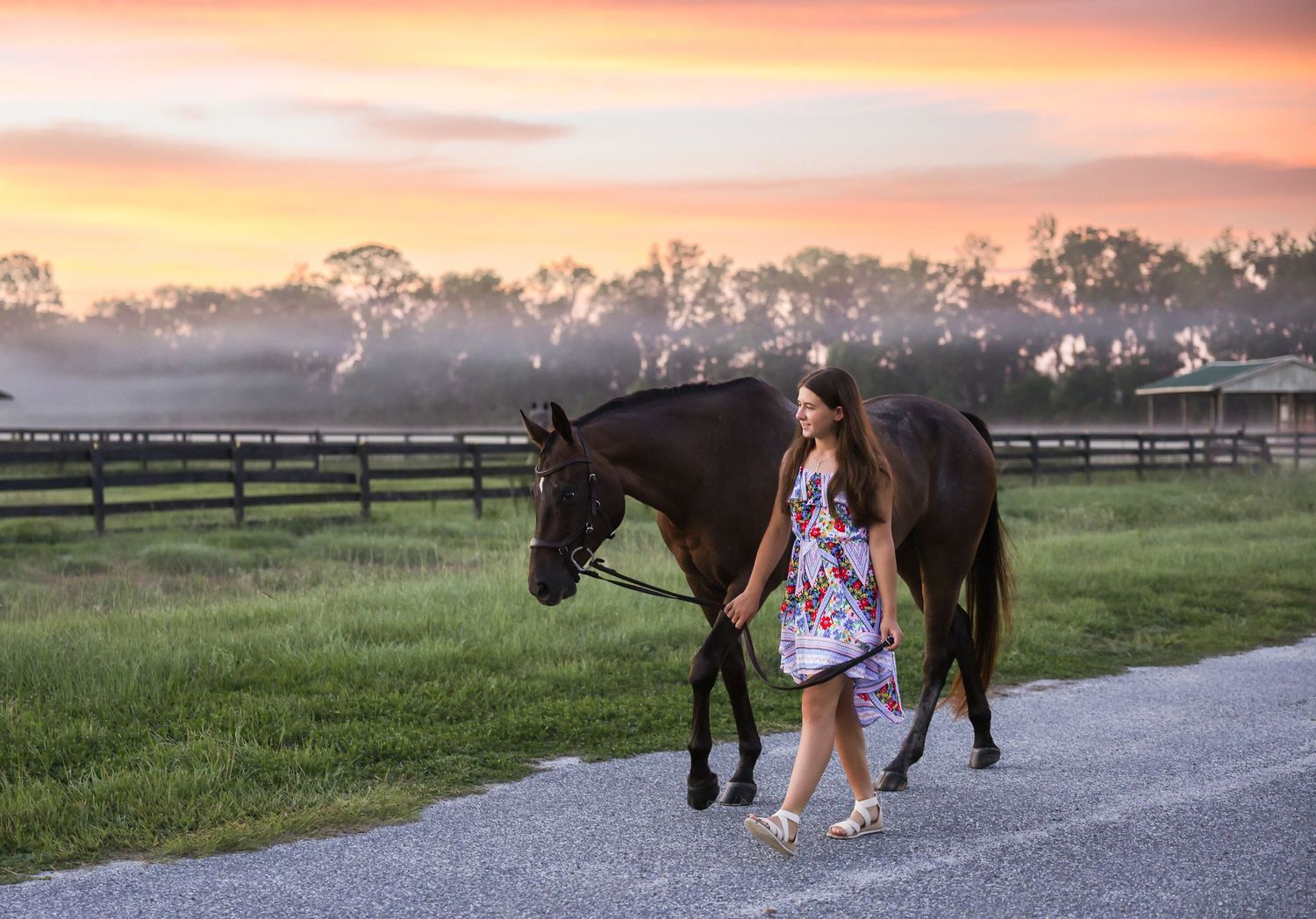 Young woman in floral dress walking dark horse at sunset on farm path with colorful sky and misty fields.