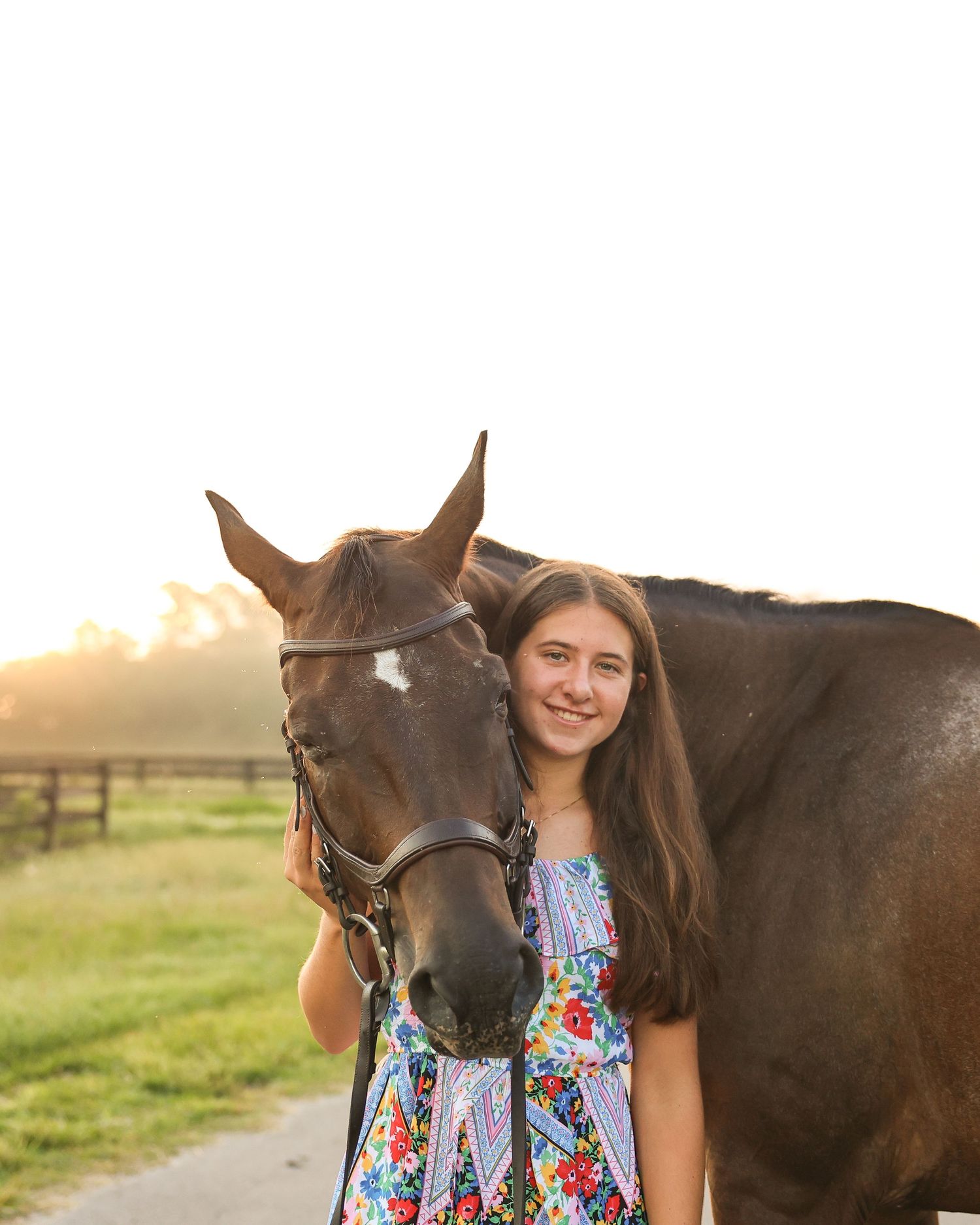 A smiling teenage girl in a floral dress poses with a dark brown horse at golden hour on a sunny farm path.