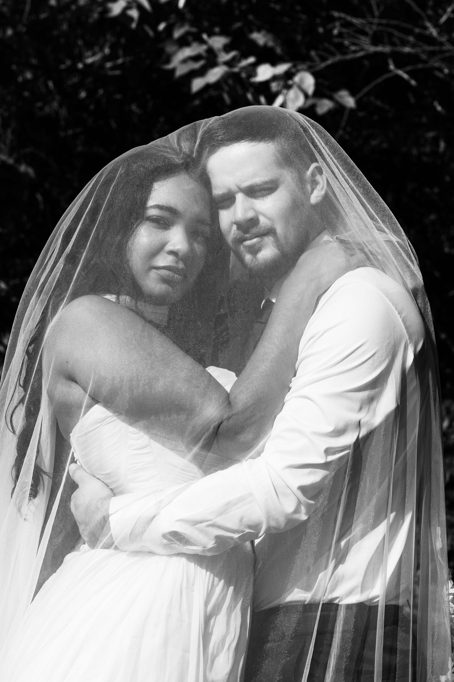 Black and white photo of a bride and groom embracing under a wedding veil outdoors.