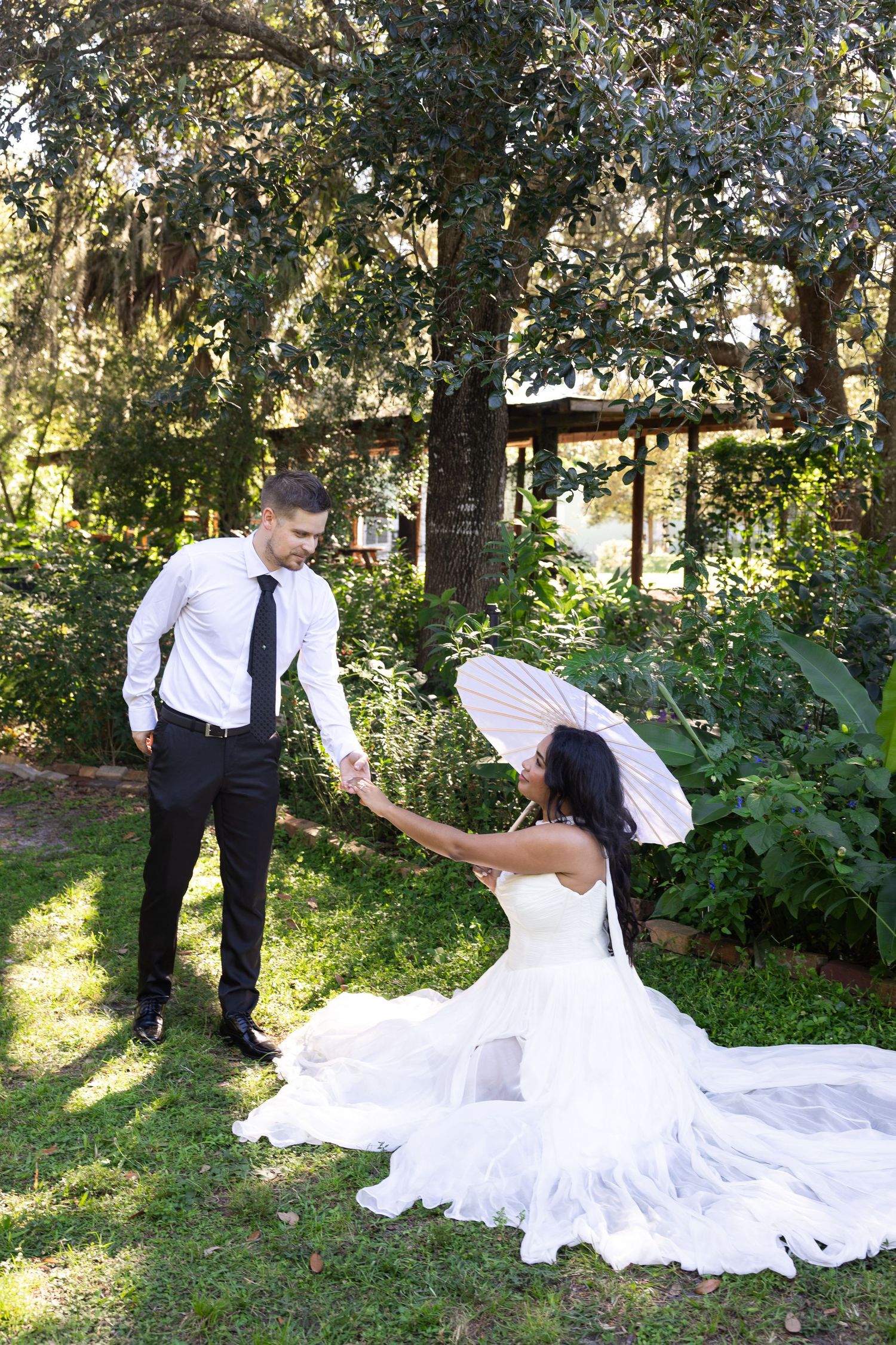 Bride in white gown holds parasol while groom in black suit assists her on lush green garden grounds.
